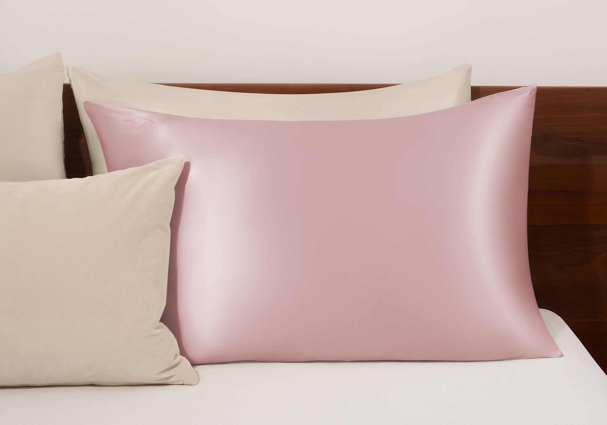 A rose pink Sleep silk pillowcase arranged on a bed with matching neutral-toned accent pillows against a wooden headboard.