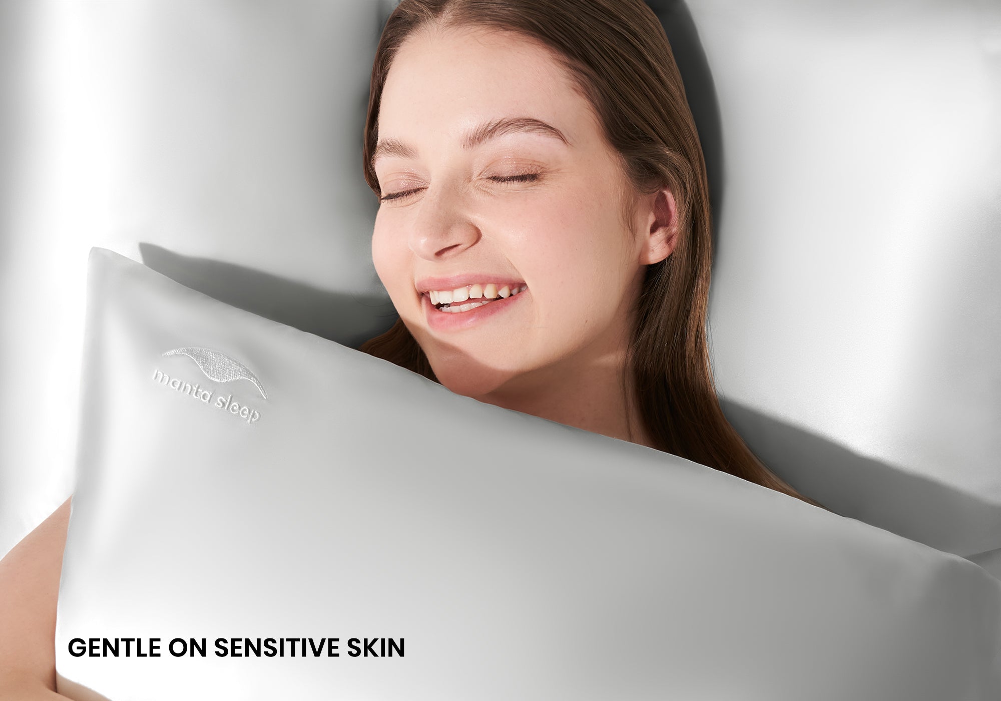 A smiling woman with closed eyes hugging a crystal silver Manta Sleep silk pillowcase. Text overlay: "Gentle on sensitive skin".