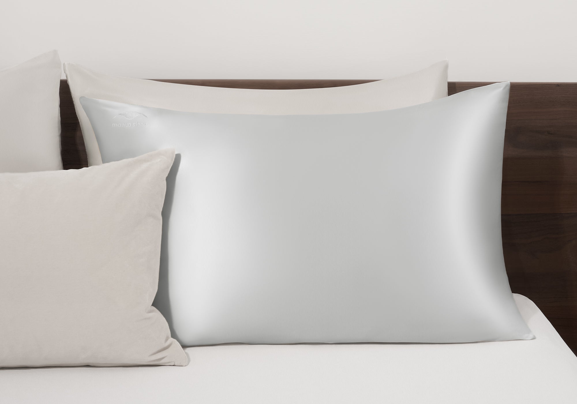 A crystal silver Manta Sleep silk pillowcase arranged on a bed with matching neutral-toned accent pillows against a wooden headboard.