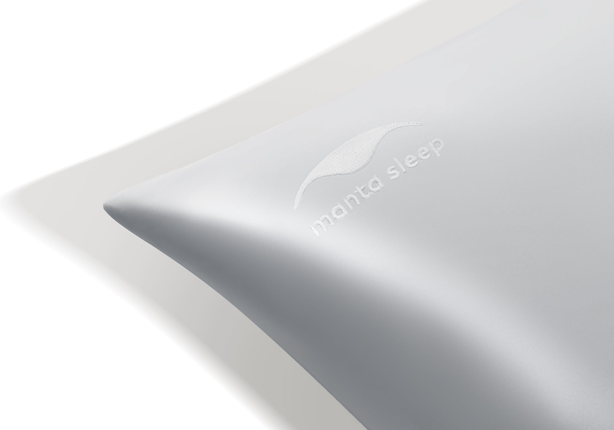 Extreme close-up of the tonal, embroidered Manta Sleep logo on the corner of a crystal silver silk pillowcase.
