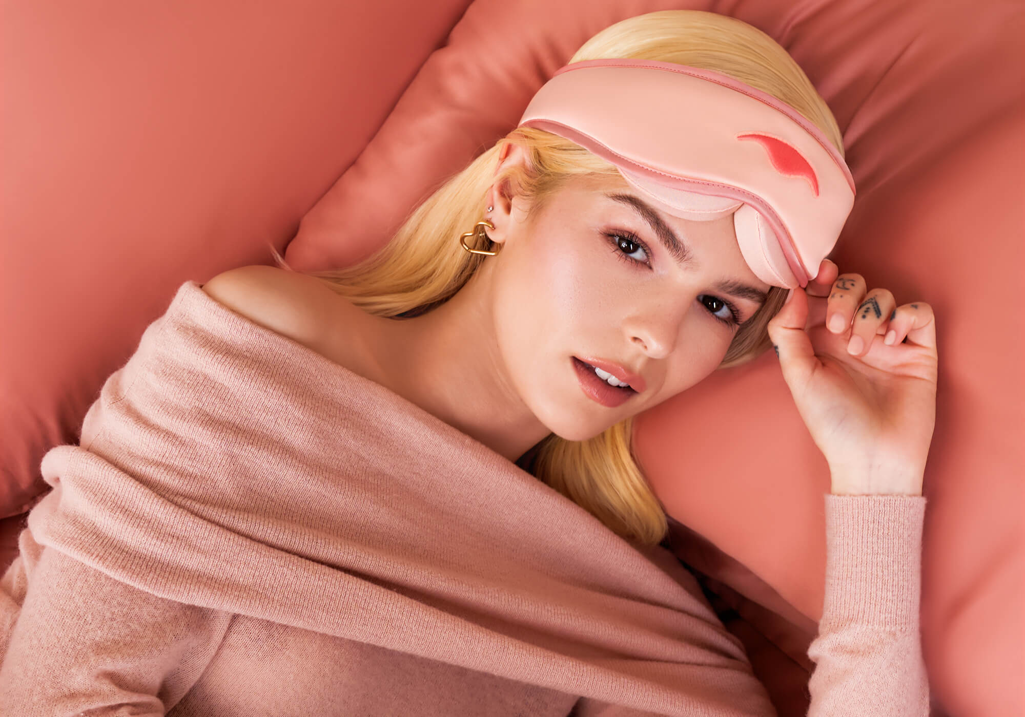 Top-down view of a blonde person lying on pink silk pillows, wearing a pink Manta SILK Sleep Mask pushed up on their forehead.