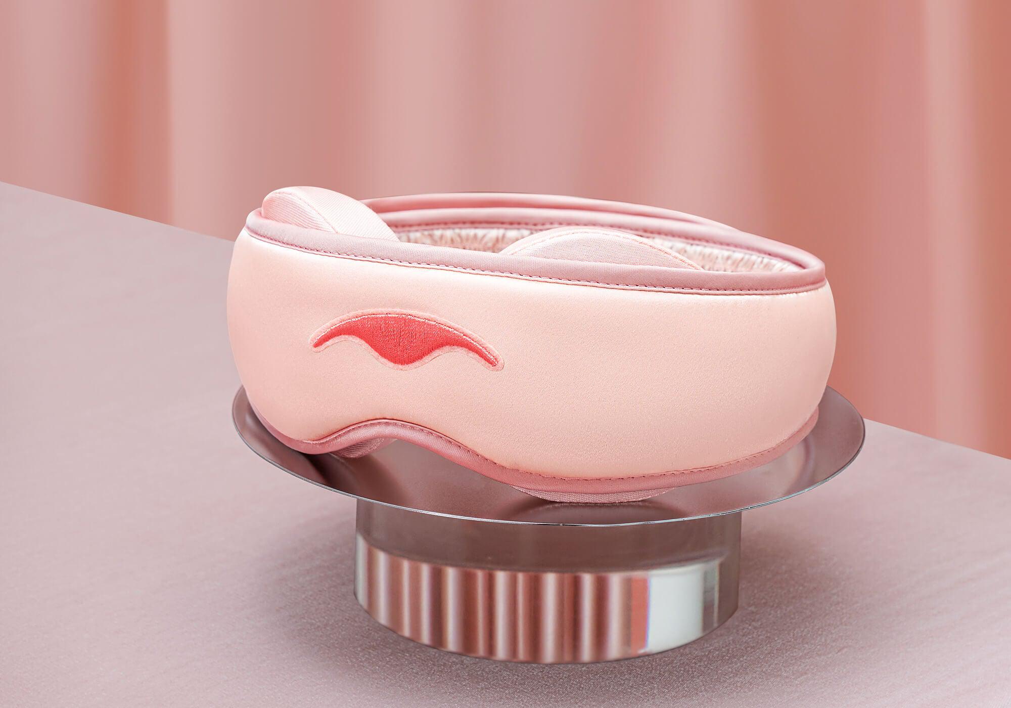 A pink Manta SILK Sleep Mask resting on a metallic silver display stand, set against a soft pink draped fabric background.