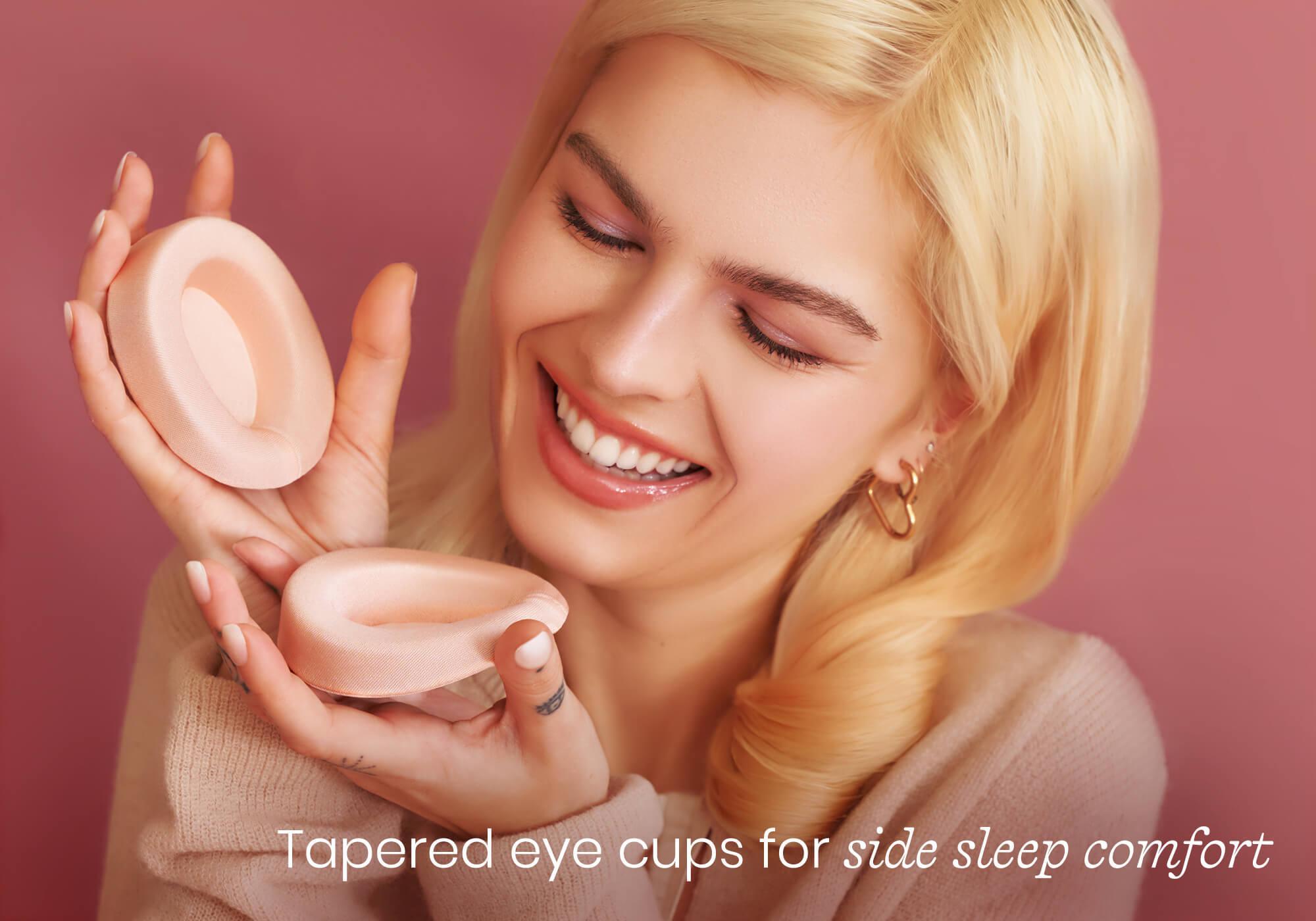 Smiling person holding two contoured pink eye cups. Text overlay reads "Tapered eye cups for side sleep comfort".