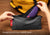 Hands placing a blue and red Manta PRO Sleep Mask into the grey zippered travel pouch, with a yellow backpack in the background. Text overlay: "Designed to fit any Manta mask".