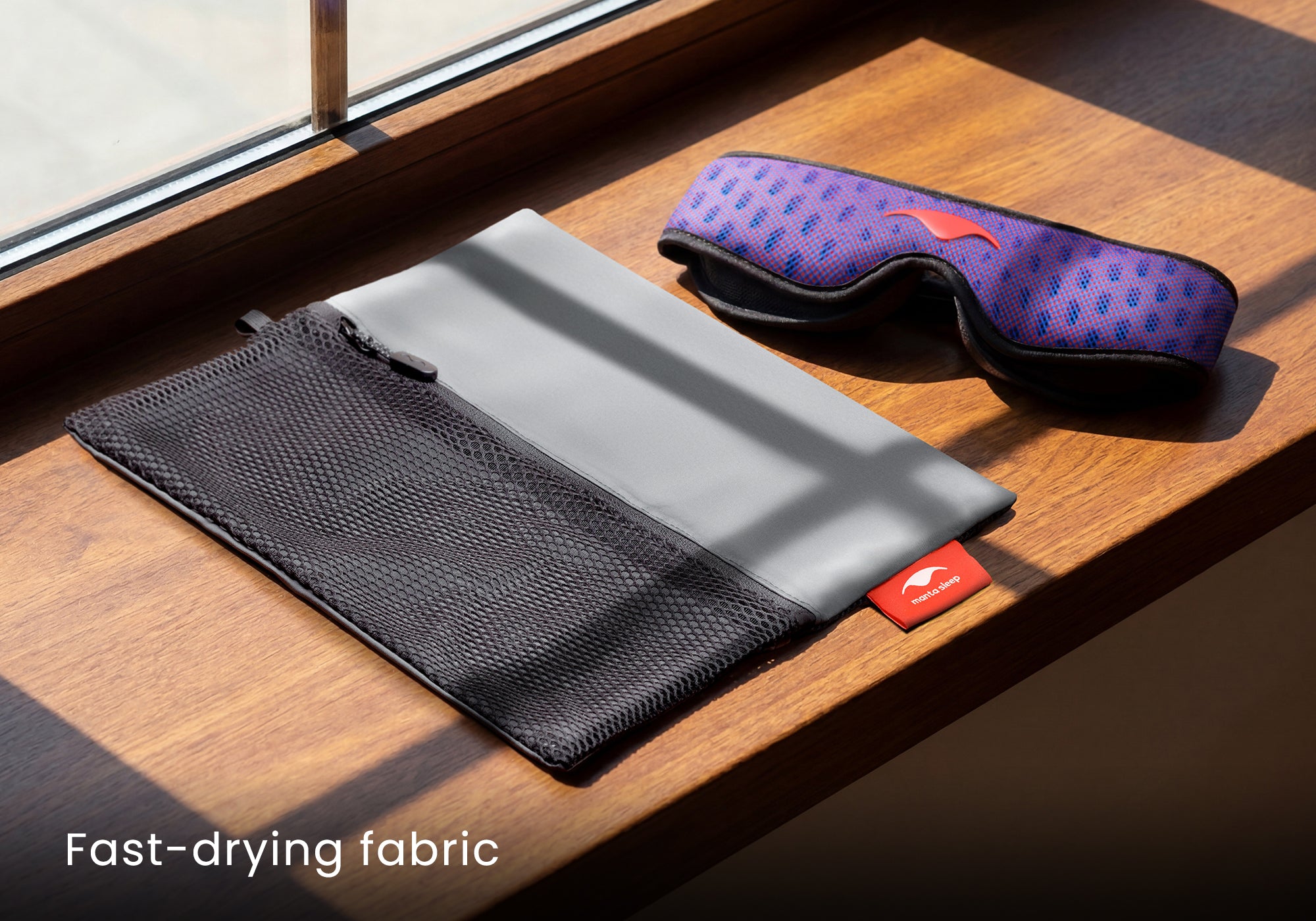 An unfolded grey and black mesh Manta Travel & Laundry Bag resting on a wooden window sill next to a blue and red Manta PRO Sleep Mask in the sunlight. Text overlay: "Fast-drying fabric".