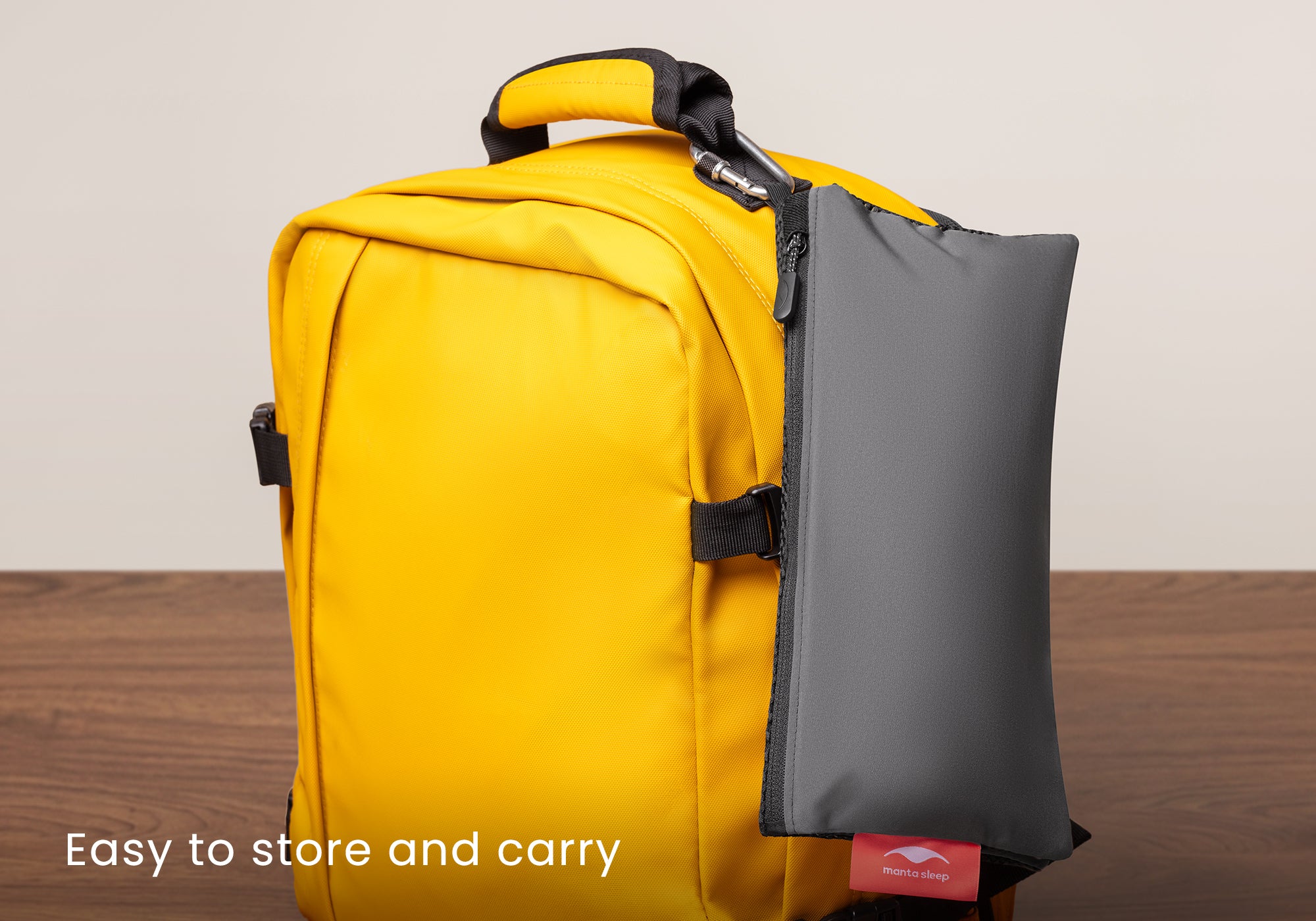 A folded grey Manta travel pouch clipped onto the side of a bright yellow travel backpack using a carabiner. Text overlay: "Easy to store and carry".