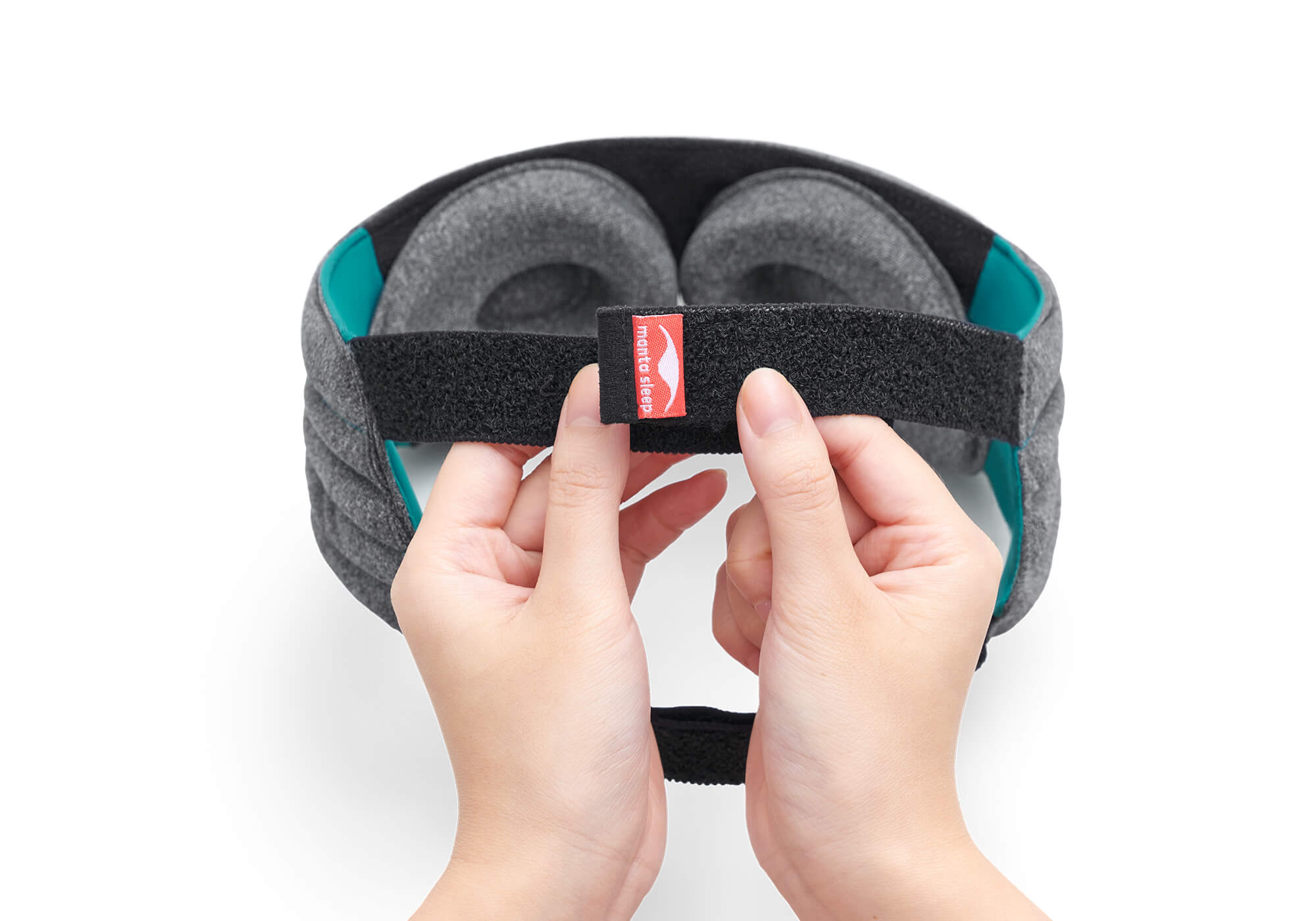 A close-up view of two hands adjusting the micro-hook velcro closure at the back of the Manta WEIGHTED Sleep Mask. The black elastic strap features a small red tag with the white "manta sleep" logo, while the heather-gray eye cups and bright teal side linings are visible in the background against a clean white surface.