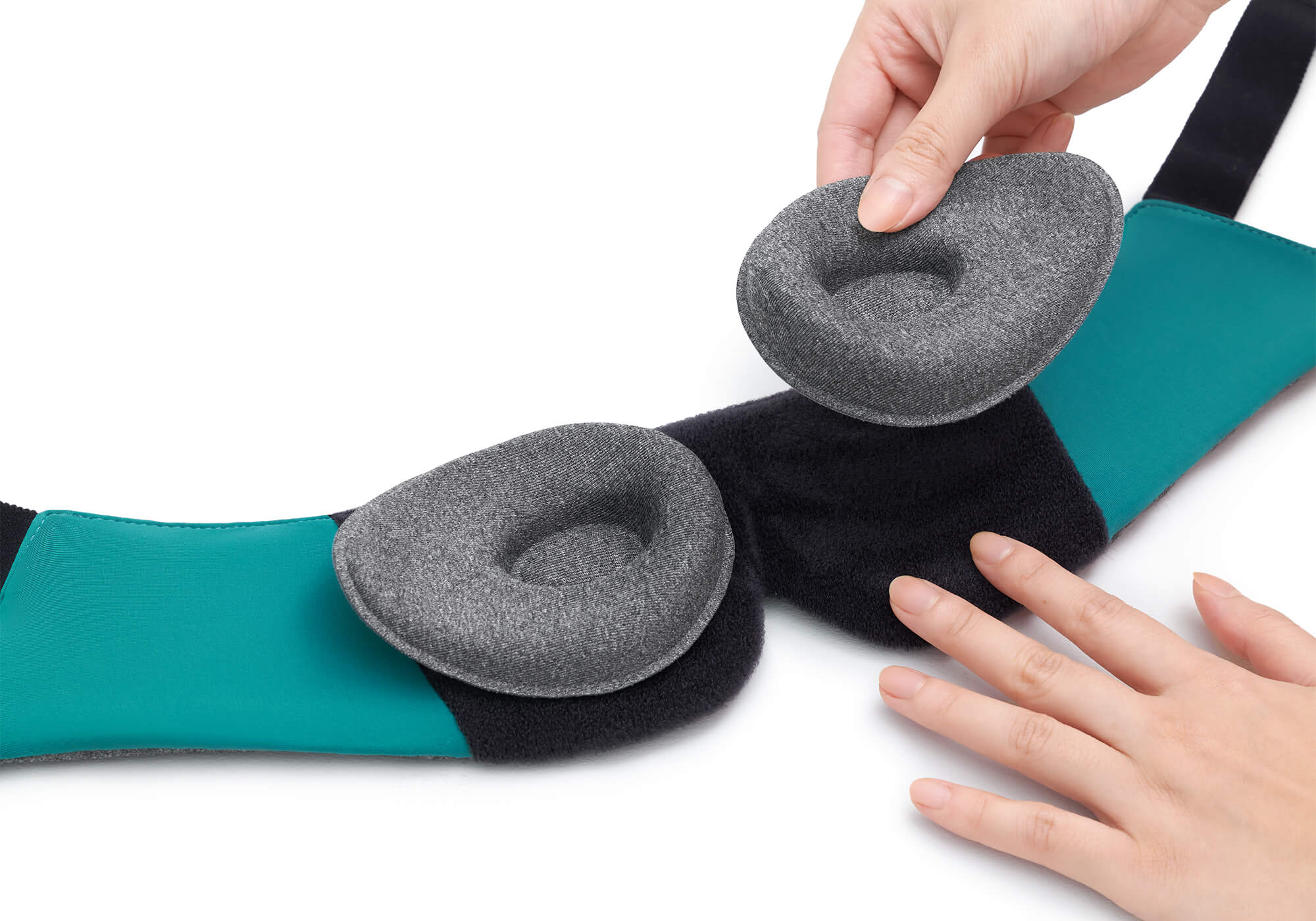 A close-up shot demonstrating the modularity of the Manta WEIGHTED Sleep Mask on a white background. One hand removes a contoured heather-gray fabric eye cup from the wide, black plush hook-and-loop track on the mask's interior, while another hand steadies the frame, showing the bright teal side lining.
