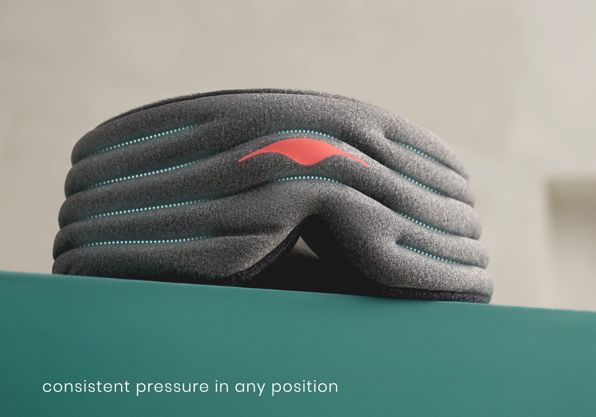 A detailed close-up shot of the front of the heather-gray Manta WEIGHTED Sleep Mask resting on a teal surface. Curved rows of glowing blue dotted lines overlay the quilted channels of the padded body to illustrate the distribution of internal weights. A coral-red Manta logo is featured in the center, and white text at the bottom left reads, "consistent pressure in any position."