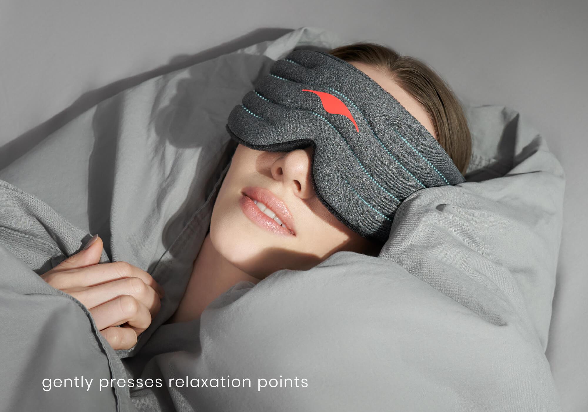 A close-up lifestyle portrait of a person lying in bed, tucked under a gray duvet and pillow. They are wearing the heather-gray Manta WEIGHTED Sleep Mask over their eyes. Glowing blue dotted lines map out the quilted rows across the mask to signify calming pressure points, and white text at the bottom left reads, "gently presses relaxation points."