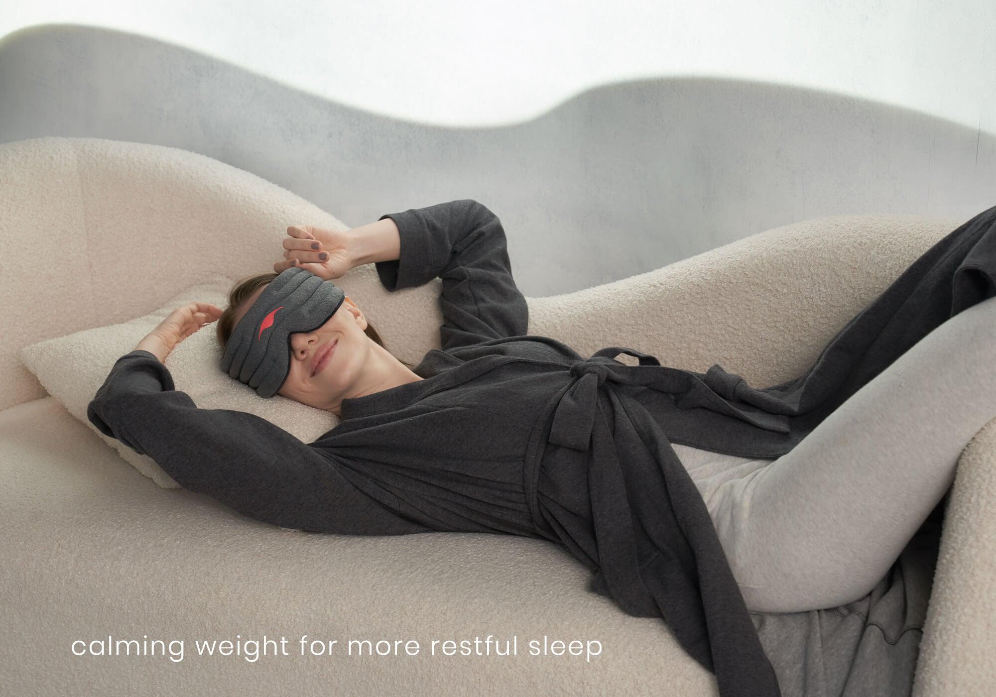 A high-angle lifestyle shot of a person lying down on a cozy, cream-colored textured sofa under soft lighting. The person wears gray loungewear and has their eyes covered with the heather-gray Manta WEIGHTED Sleep Mask, resting their head on a matching pillow with arms relaxed overhead. White text at the bottom left reads, "calming weight for more restful sleep."