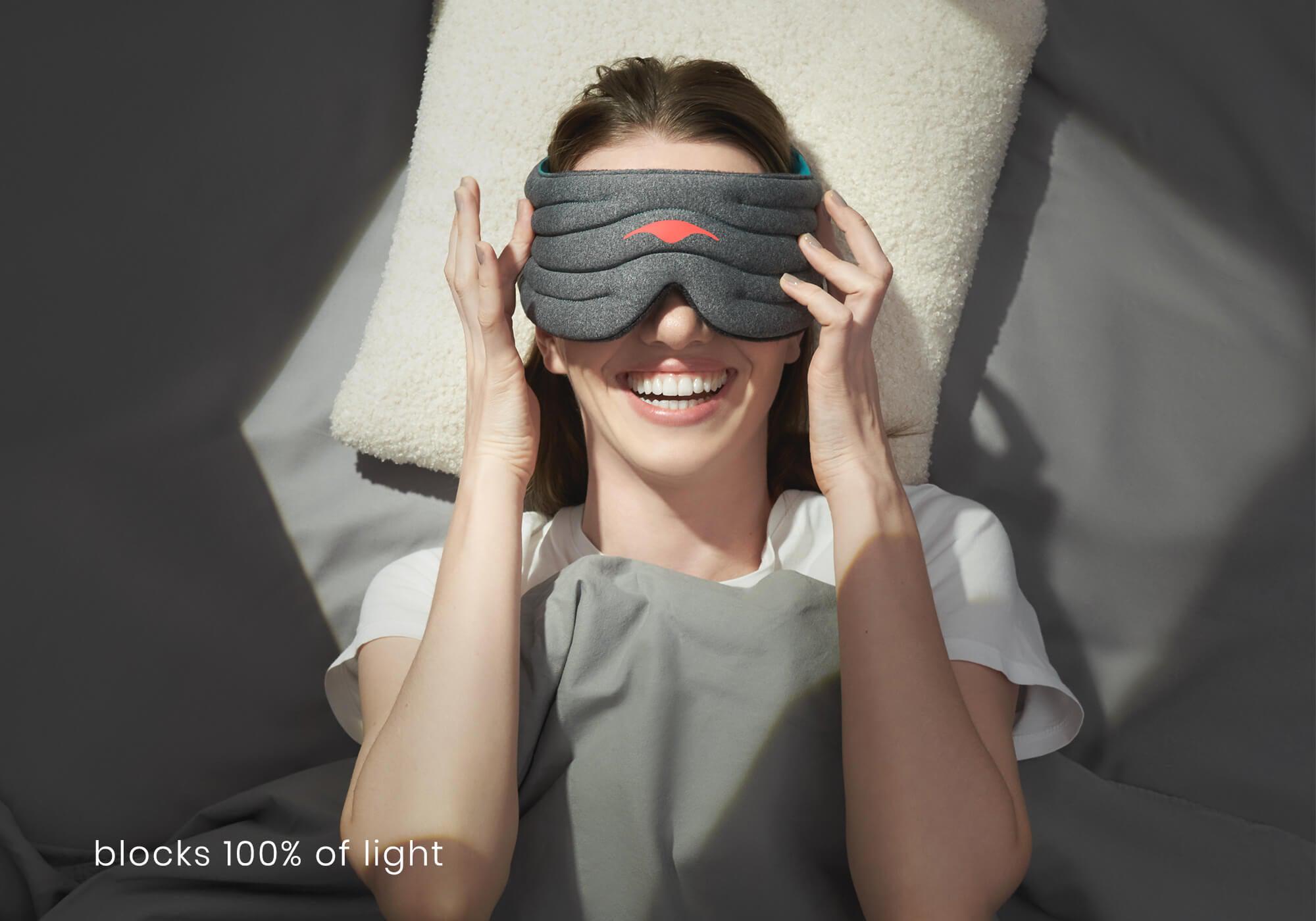 A high-angle portrait of a person lying on their back in bed, resting their head on an off-white textured pillow. They are smiling brightly while using both hands to adjust the heather-gray Manta WEIGHTED Sleep Mask over their eyes. White text at the bottom left reads, "blocks 100% of light."