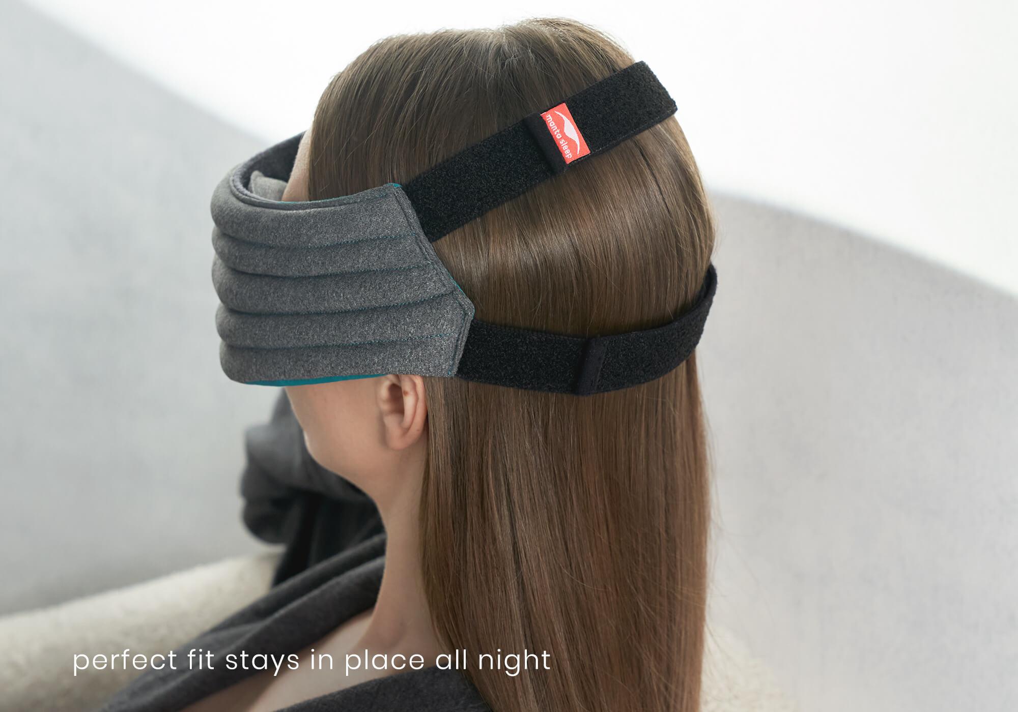 A side-profile view of a person with long brown hair wearing the heather-gray Manta WEIGHTED Sleep Mask. The shot highlights the fit of the two black elastic head straps looping securely around the back of the head, with the top strap featuring a small red brand tag. White text at the bottom left reads, "perfect fit stays in place all night."