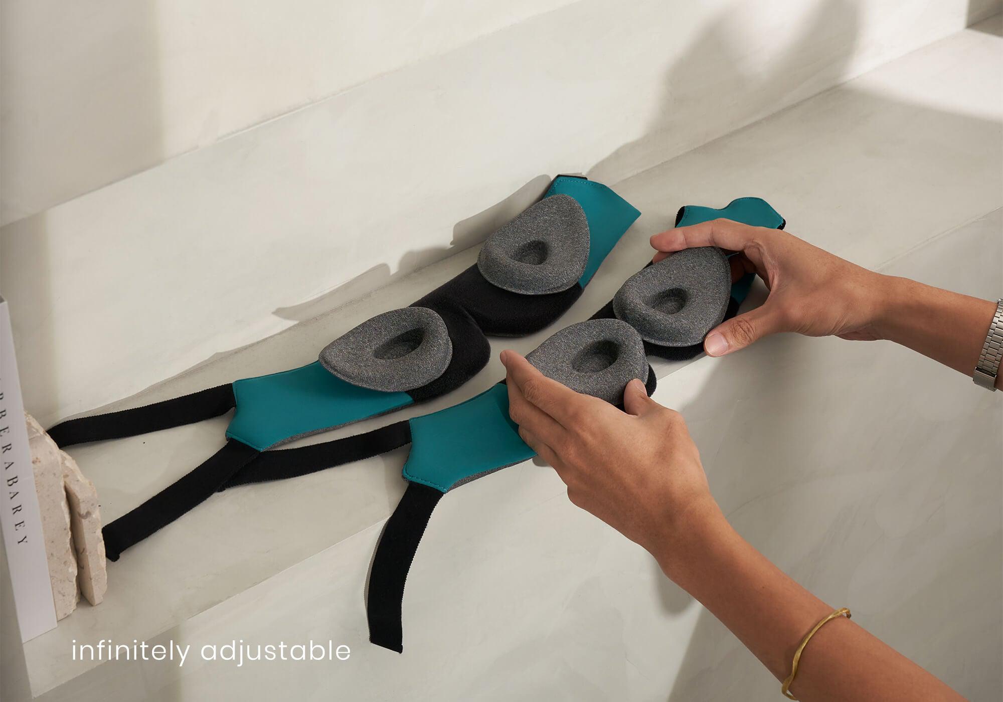A lifestyle product shot showing a pair of hands assembling the Manta WEIGHTED Sleep Mask on a stone shelf. The main frame is laid flat to reveal its bright teal inner lining and black hook-and-loop track, while the hands position two matching heather-gray plush eye cups onto the band. White text at the bottom left reads, "infinitely adjustable."