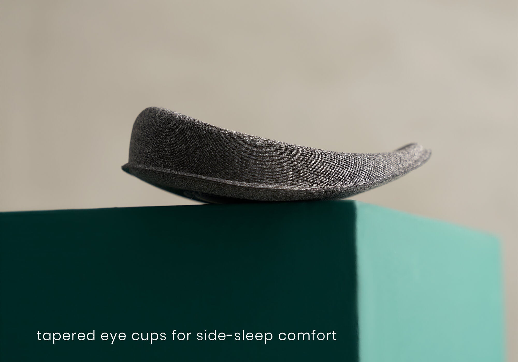 A detailed side-profile product shot of a single heather-gray eye cup resting on a teal box. The image highlights the distinctive curved and tapered edge profile designed to minimize bulk for side sleepers. White text at the bottom left reads, "tapered eye cups for side-sleep comfort."