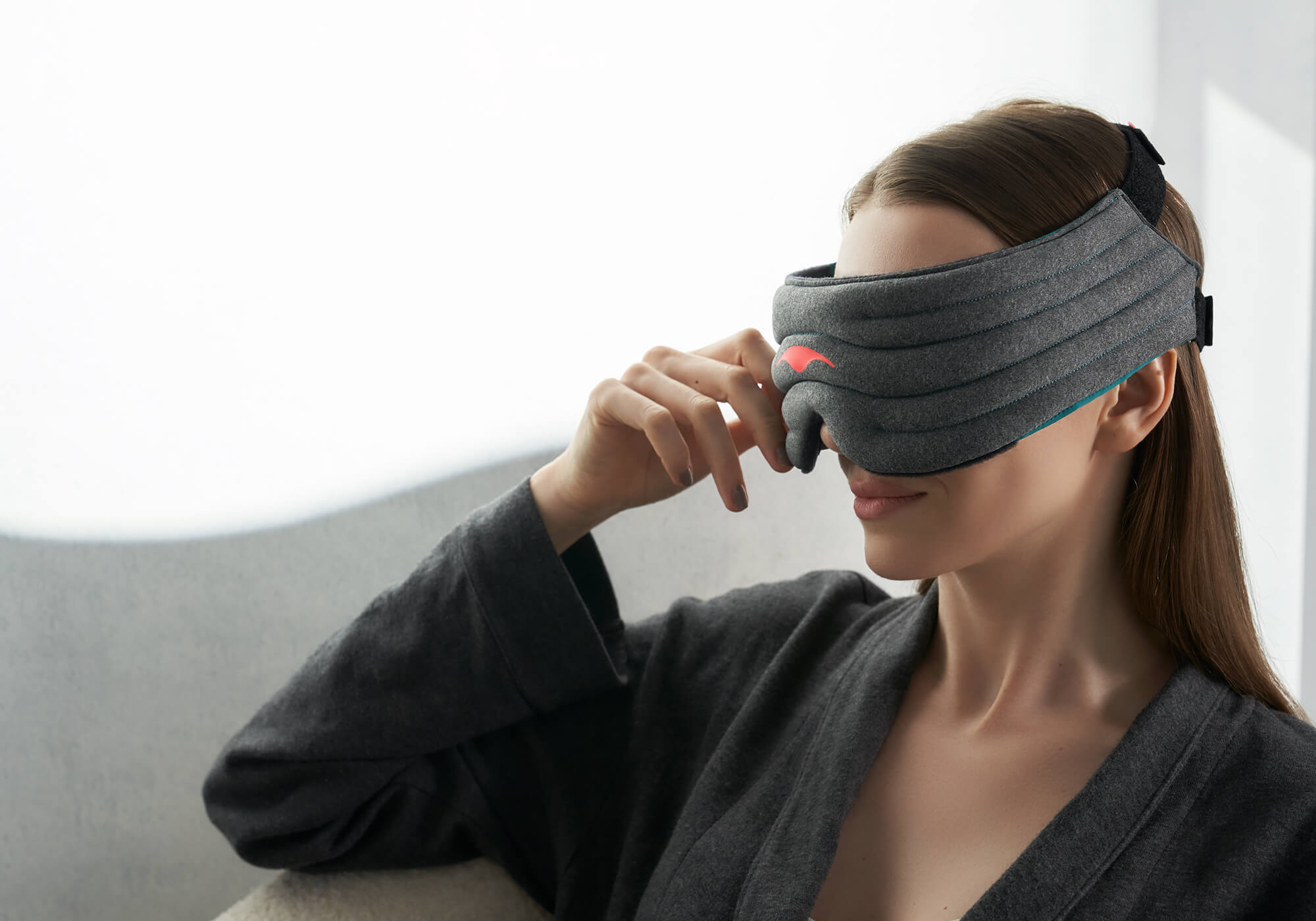 An angled close-up portrait of a person sitting in a dimly lit room wearing a dark gray robe. They are wearing the heather-gray Manta WEIGHTED Sleep Mask, resting a hand near the side of the band. The mask fits snugly against their face, displaying its segmented, quilted rows and a red logo.