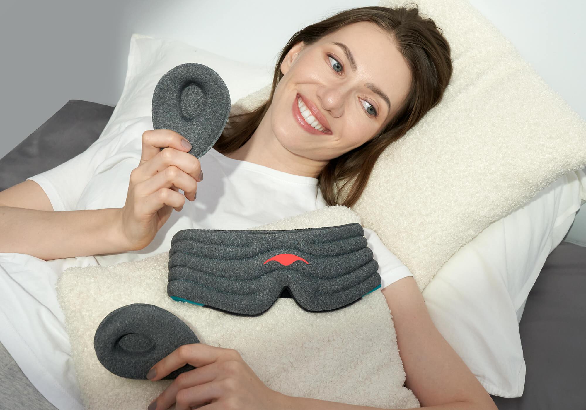 A high-angle portrait of a smiling person with brown hair resting in bed on a white pillow. They are holding up the two detached heather-gray plush eye cups, while the main body of the Manta WEIGHTED Sleep Mask rests on the cream-colored blanket in front of them.