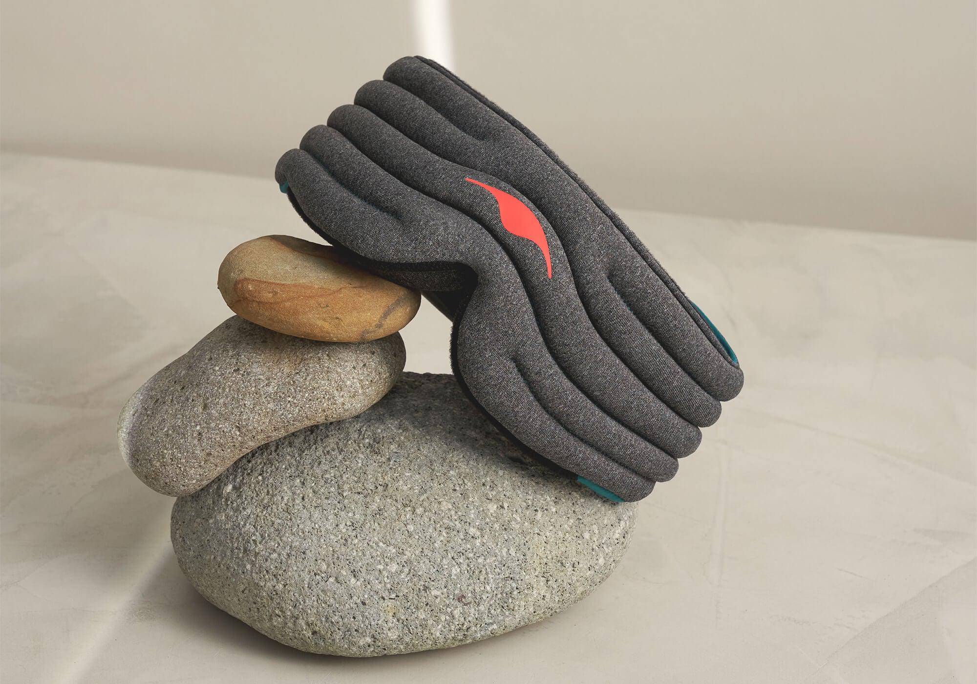 A creative product shot featuring the heather-gray Manta WEIGHTED Sleep Mask balanced gracefully atop a small stack of three smooth, natural river stones. The scene is illuminated by soft light on a neutral surface, showcasing the quilted rows of the mask and its red emblem.