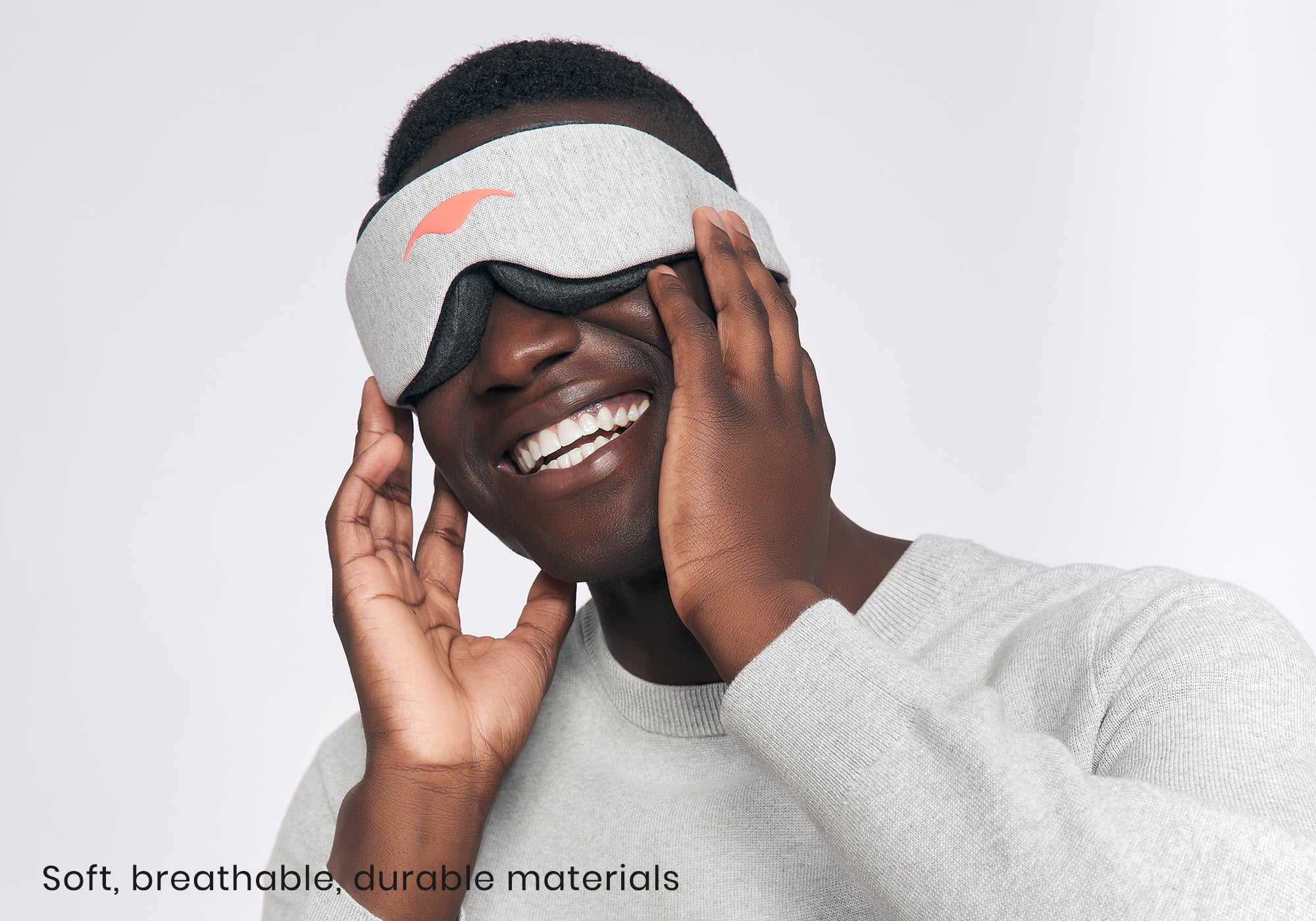 A smiling person touches the sides of a heather grey Manta Sleep Mask Classic. Text reads "Soft, breathable, durable materials".