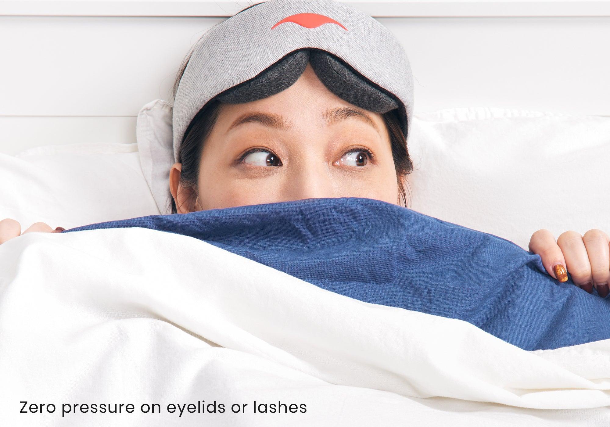 A person peeking out from under a blue and white blanket while wearing a grey sleep mask. Text reads "Zero pressure on eyelids or lashes".