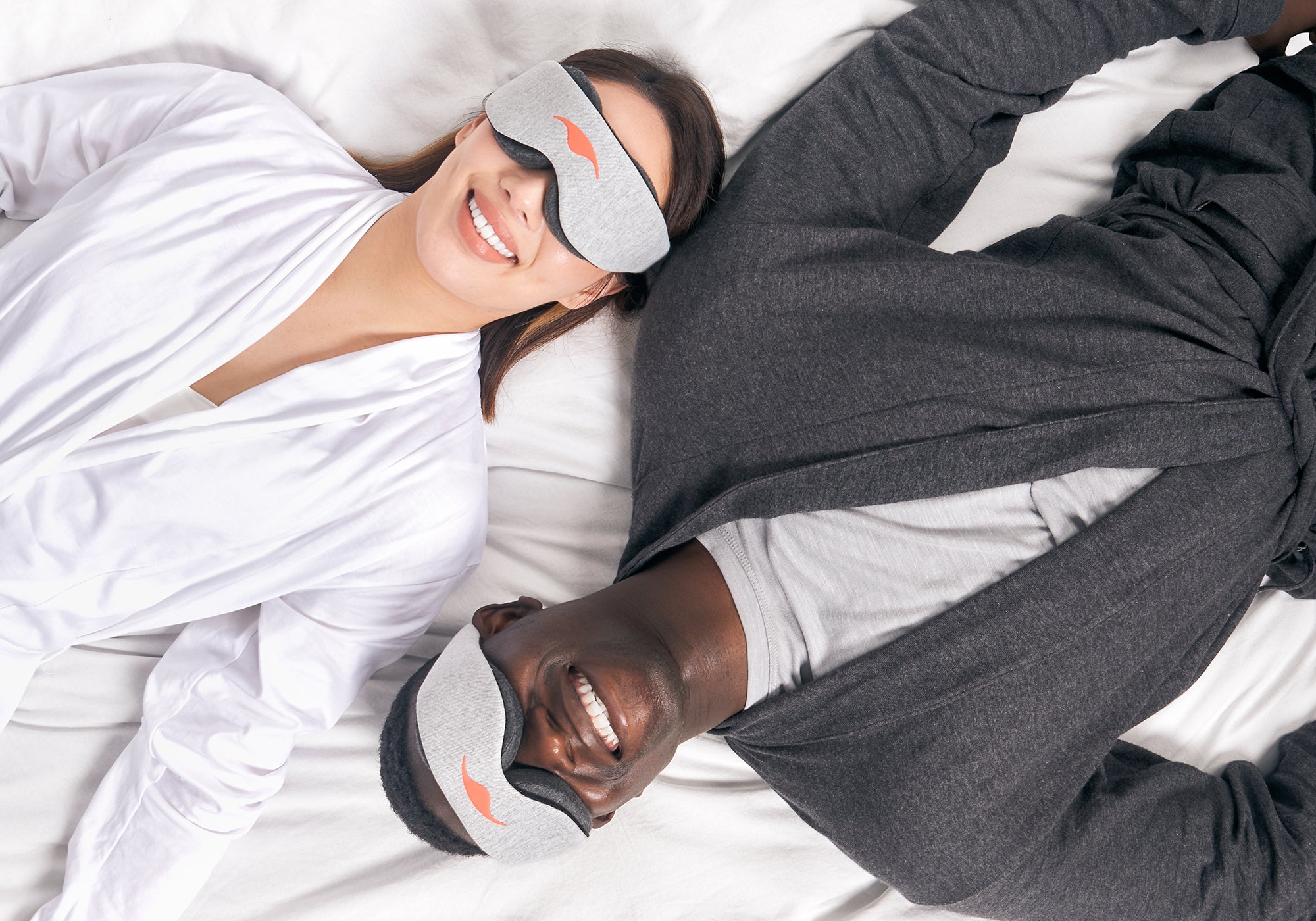 Overhead view of two smiling people lying side-by-side in bed, both wearing heather grey Manta Sleep Mask Classic units.