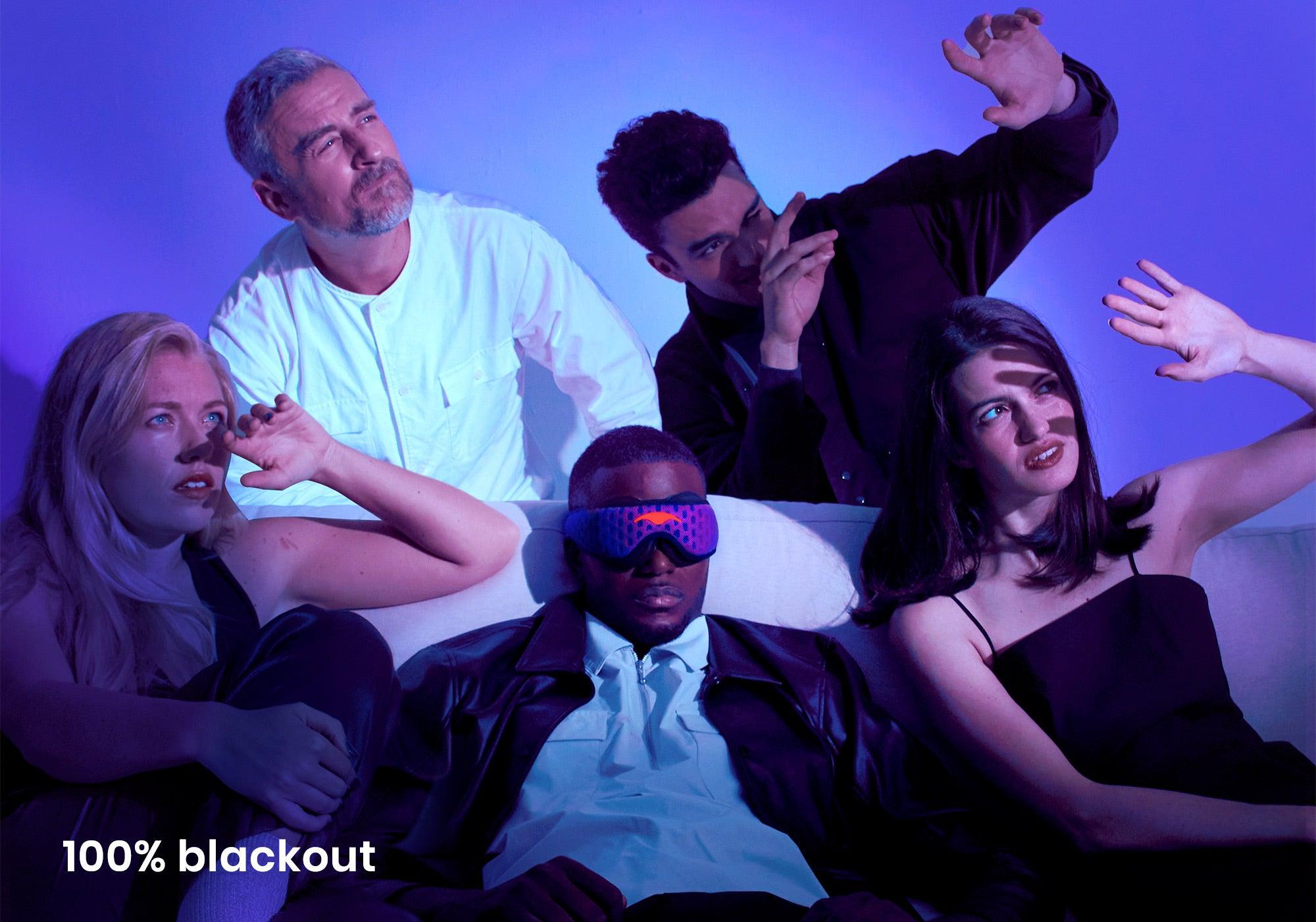 Five people on a couch under harsh blue light shields their eyes from glare, except one man wearing a black-out Manta Sleep Mask PRO, with caption "100% blackout".