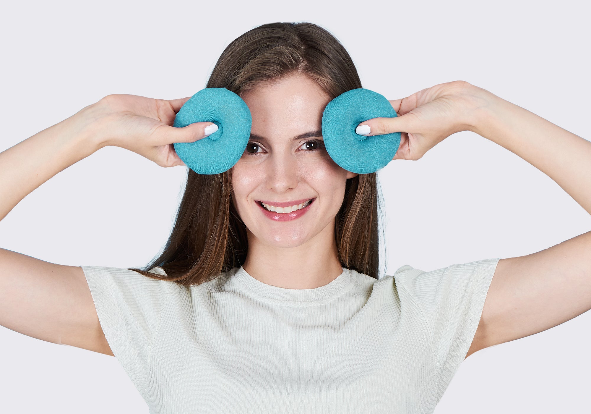 A smiling woman holding two round, blue-teal fabric eye cups over her eyes against a plain light gray background.