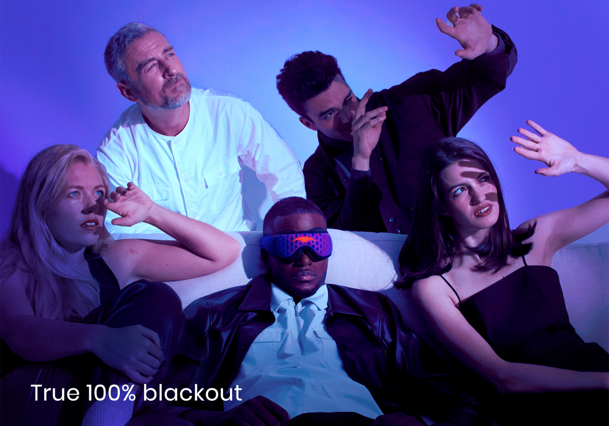 Five people on a couch under harsh blue light shields their eyes from glare, except one man wearing a black-out Manta PRO Sleep Mask, featuring text 'True 100% blackout'.