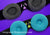 Two black Manta sleep mask head straps, one fitted with gray eye cups and purple patch holder, and one with blue eye cups and green patch holder.
