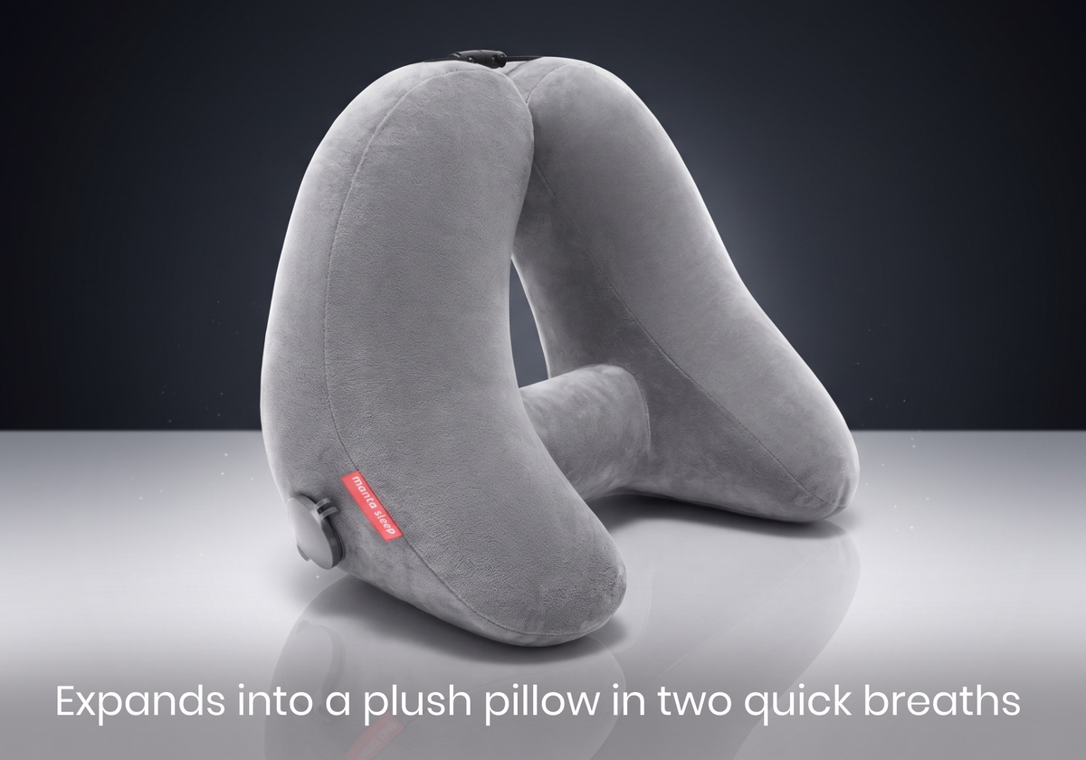 A gray inflatable travel neck pillow shown in its inflated U-shape on a reflective surface with a dark gradient background. White text at the bottom reads 'Expands into a plush pillow in two quick breaths'.