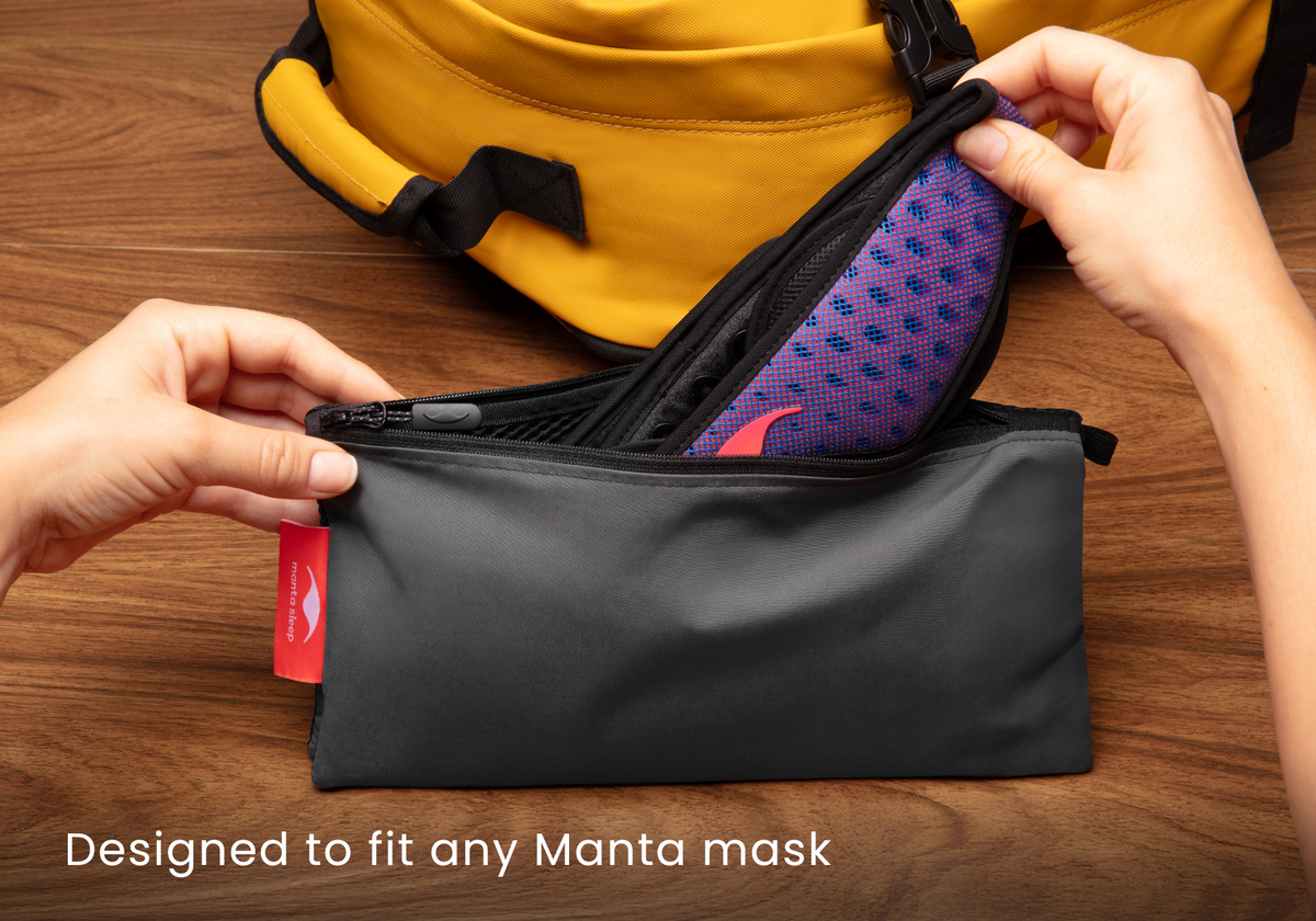 Hands placing a blue and red Manta PRO Sleep Mask into the grey zippered travel pouch, with a yellow backpack in the background. Text overlay: "Designed to fit any Manta mask".