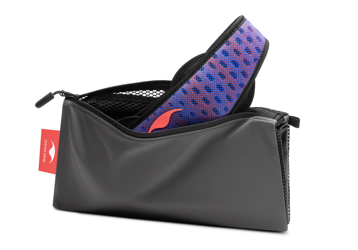 Studio shot of an open, charcoal grey zippered Manta Travel & Laundry Bag with a blue and red Manta PRO Sleep Mask peeking out, isolated on a white background.