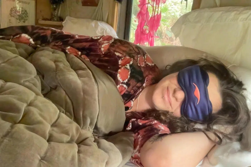 Person lying on a bed with a purple Manta SILK Sleep Mask on, wearing a floral robe.