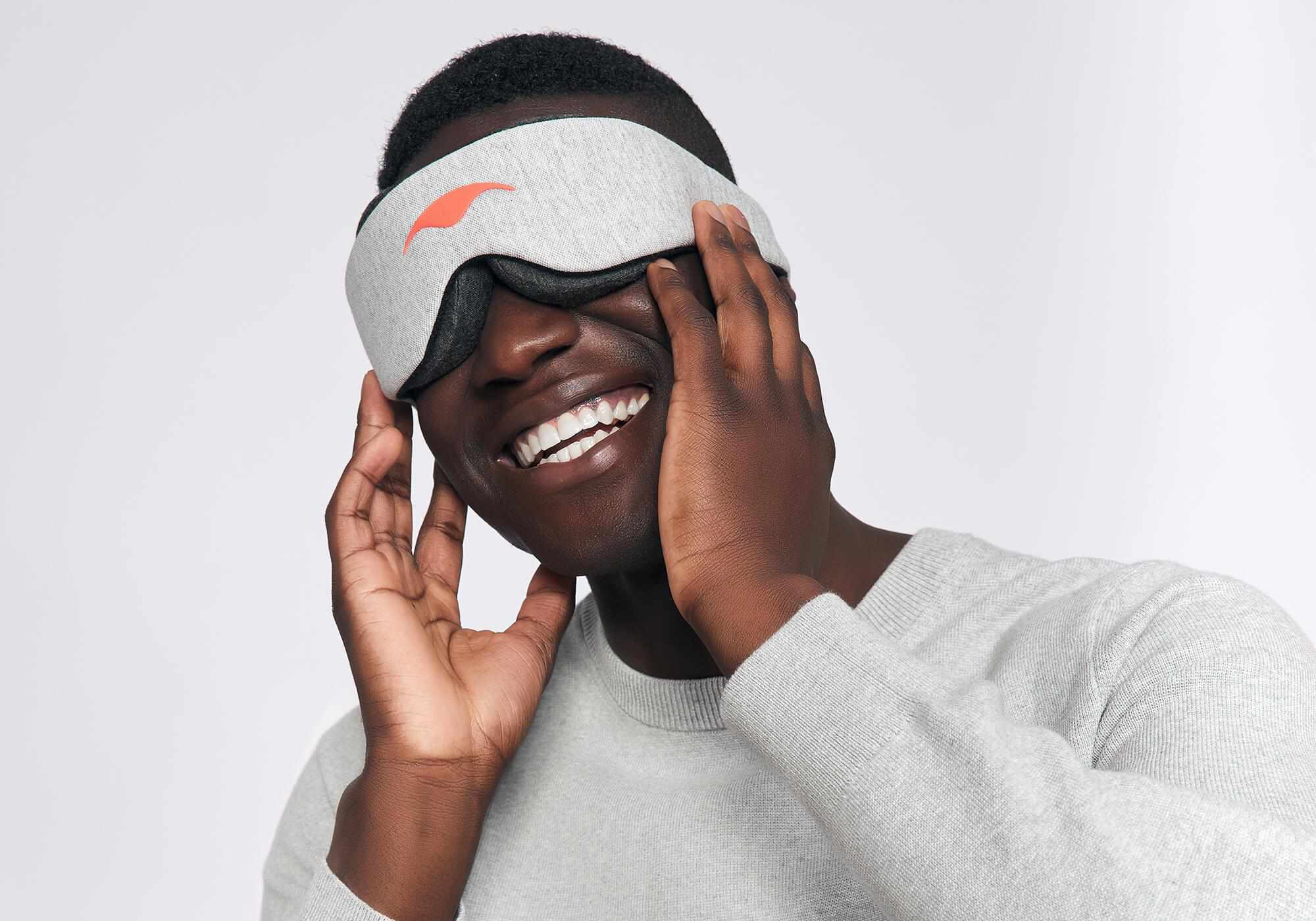 A smiling person touches the sides of a heather grey Manta Sleep Mask Classic.