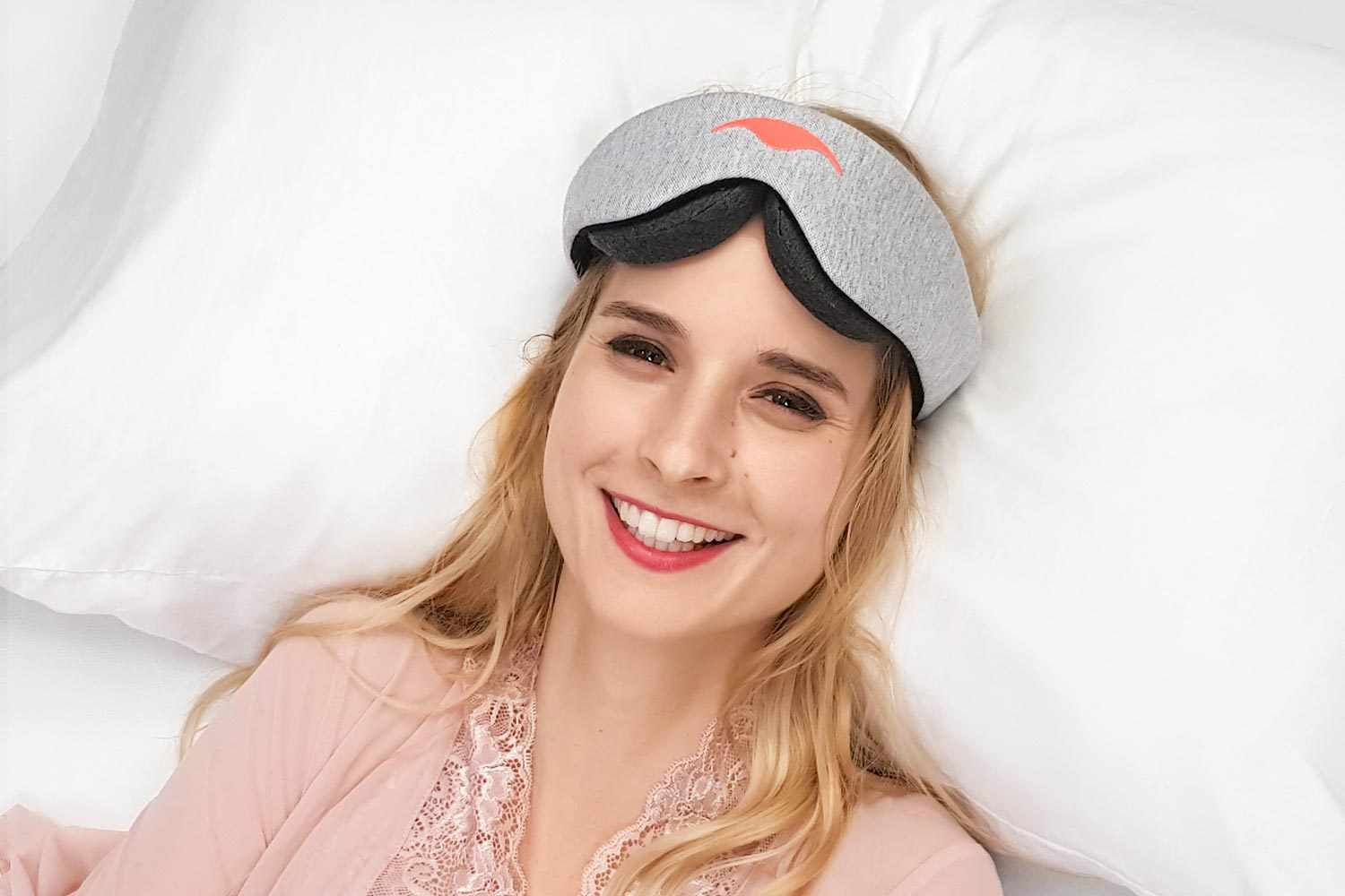 A smiling woman with blonde hair lies on a plush white pillow, wearing a light gray heather fabric Manta Sleep Mask. She is wearing a soft pink lace-trimmed robe, and a coral-colored manta ray logo is visible on the front center of the eye mask.