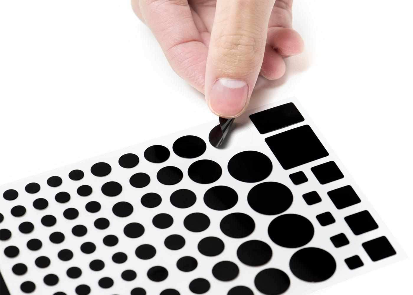 Close-up view of a hand peeling a circular black blackout sticker off a sheet containing multiple rows of various sized stickers.