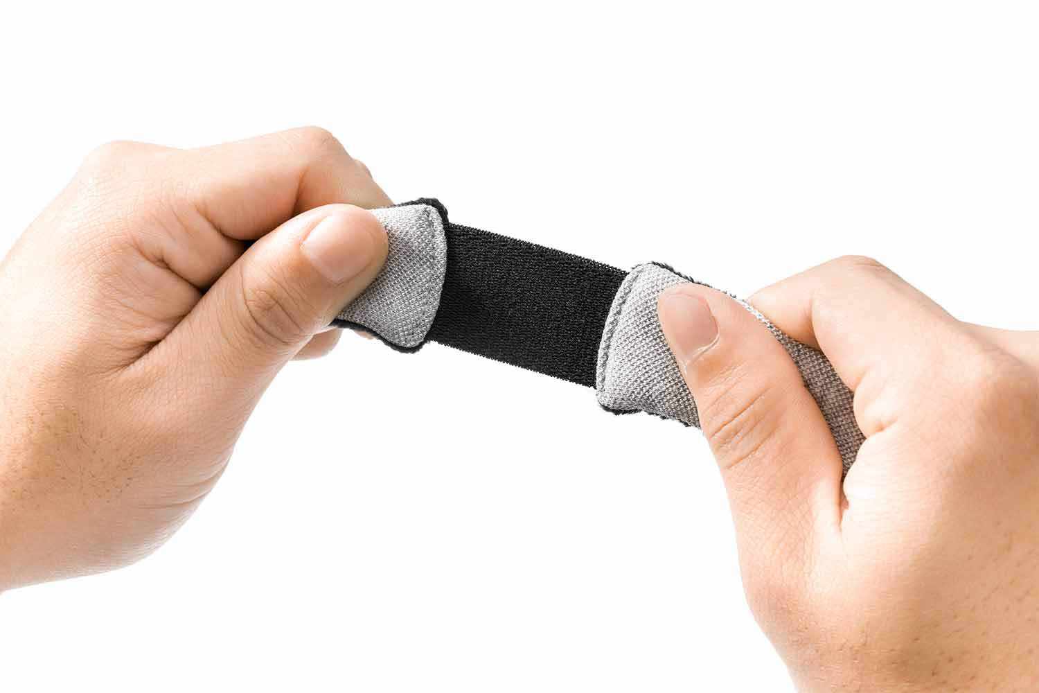 A close-up view of two hands stretching a thick, elasticated black band that connects the light gray fabric panels of the sleep mask, demonstrating the flexibility and elasticity of the head strap against a plain white background.