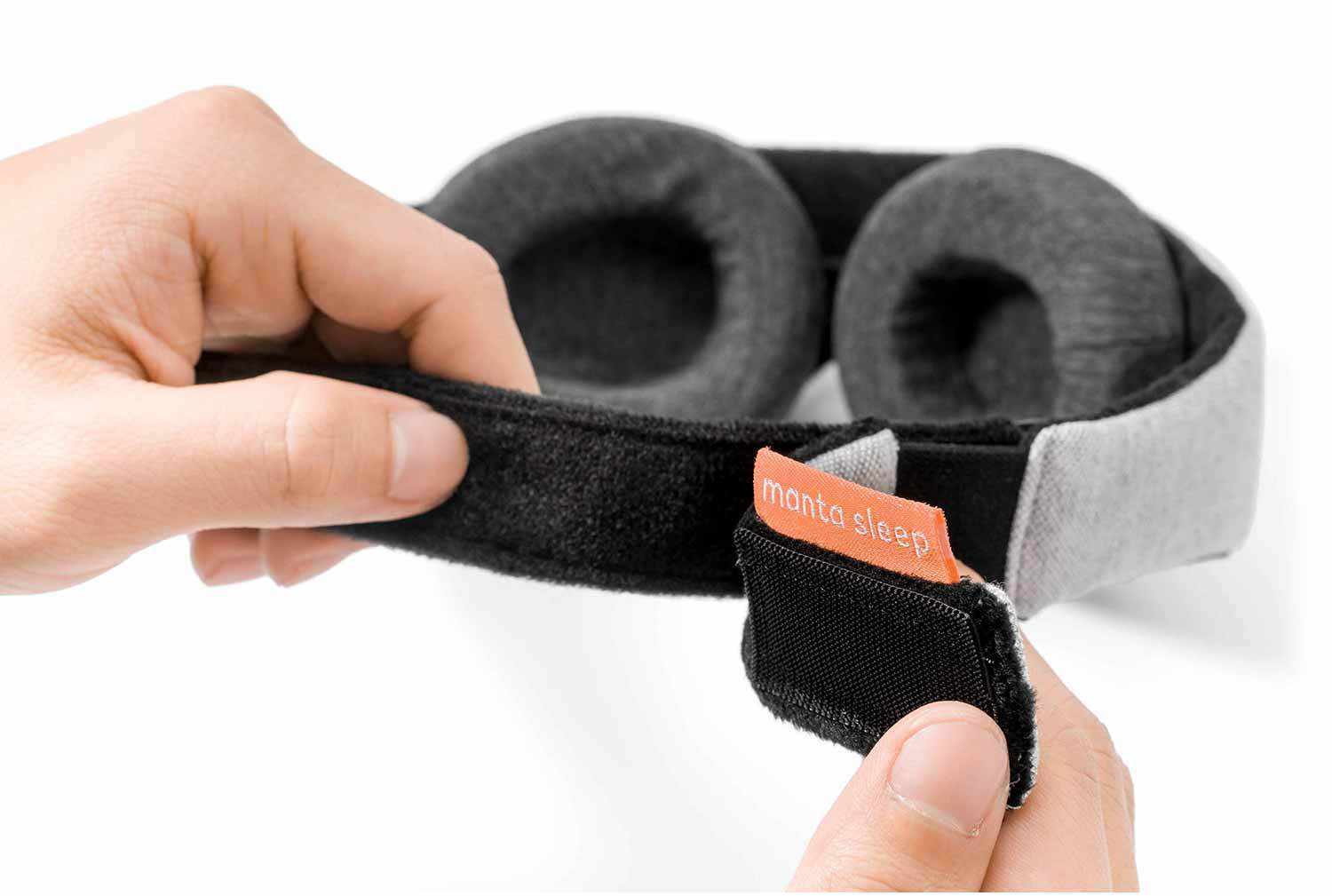 A close-up shot showing a pair of hands adjusting the black hook-and-loop fastener strap of the Manta Sleep Mask. A small, bright coral-red fabric tab featuring the white "manta sleep" text logo is prominently displayed at the closure point, with the dark gray plush eyecups visible in the background against a white backdrop.