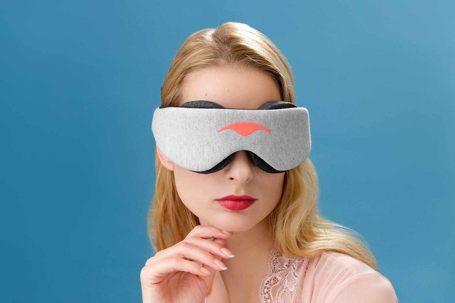 A close-up studio portrait of a blonde woman wearing a light gray Manta Sleep Mask that fully covers her eyes against a solid blue background. She is wearing a pale pink lace top and has one hand resting elegantly beneath her chin, showing red lipstick.