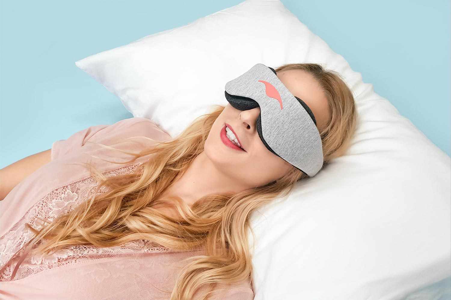 Person lying down on a fluffy white pillow, smiling while wearing a heather grey Manta Sleep Mask Classic.