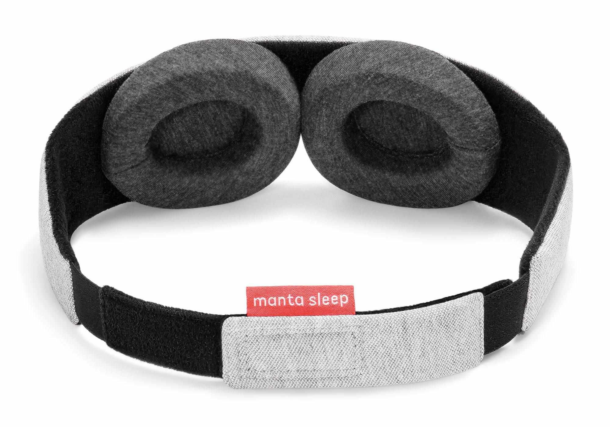 Inside view of the Manta Sleep Mask isolated on a white background, highlighting two adjustable, deep-set, circular eyecups made of dark gray heathered fabric. The black interior lining of the strap is visible, along with the adjustable light gray hook-and-loop rear closure featuring a coral-red "manta sleep" tag.