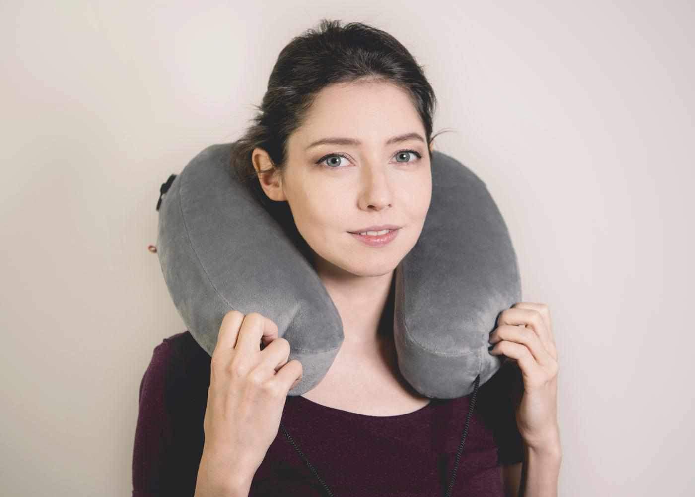 A smiling woman wearing a grey, U-shaped plush Manta Travel Pillow around her neck against a plain background.
