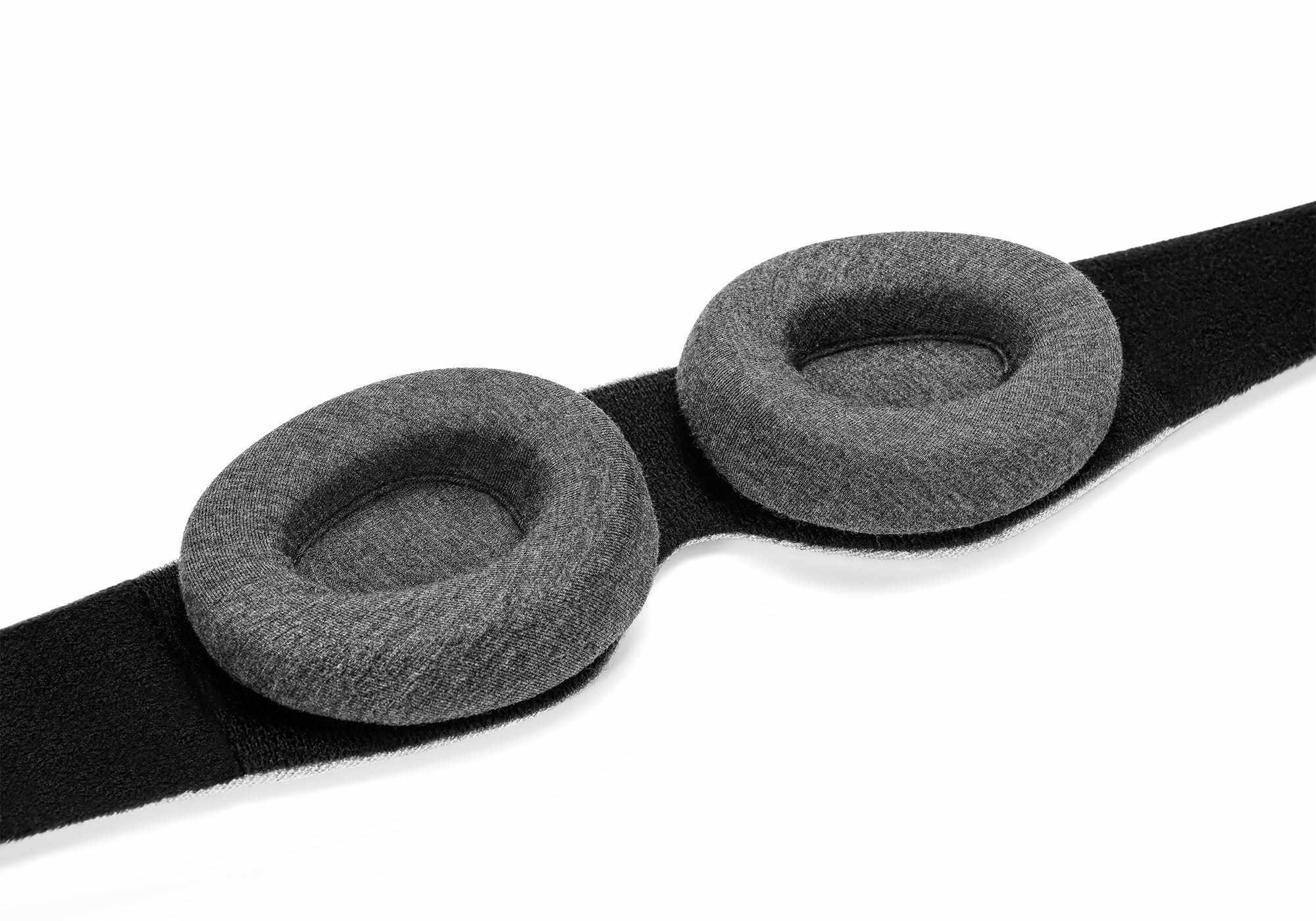 A close-up, high-angle view of the interior side of the Manta Sleep Mask laid flat on a white surface. The focus is on the two thick, cushioned, and contoured dark gray eyecups attached to a wide, soft black fabric band.