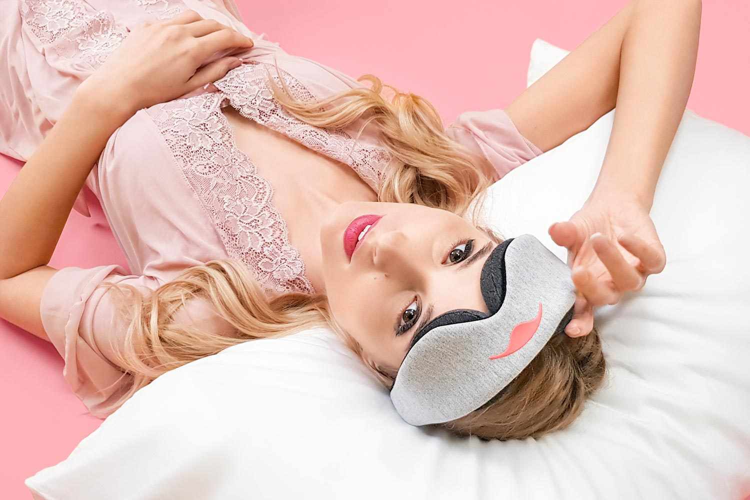 An overhead, high-angle shot of a blonde woman resting on a white pillow against a pink background. She has the light gray Manta Sleep Mask pushed up onto her forehead, looking up toward the camera with a slight smile while wearing a pink lace nightgown.
