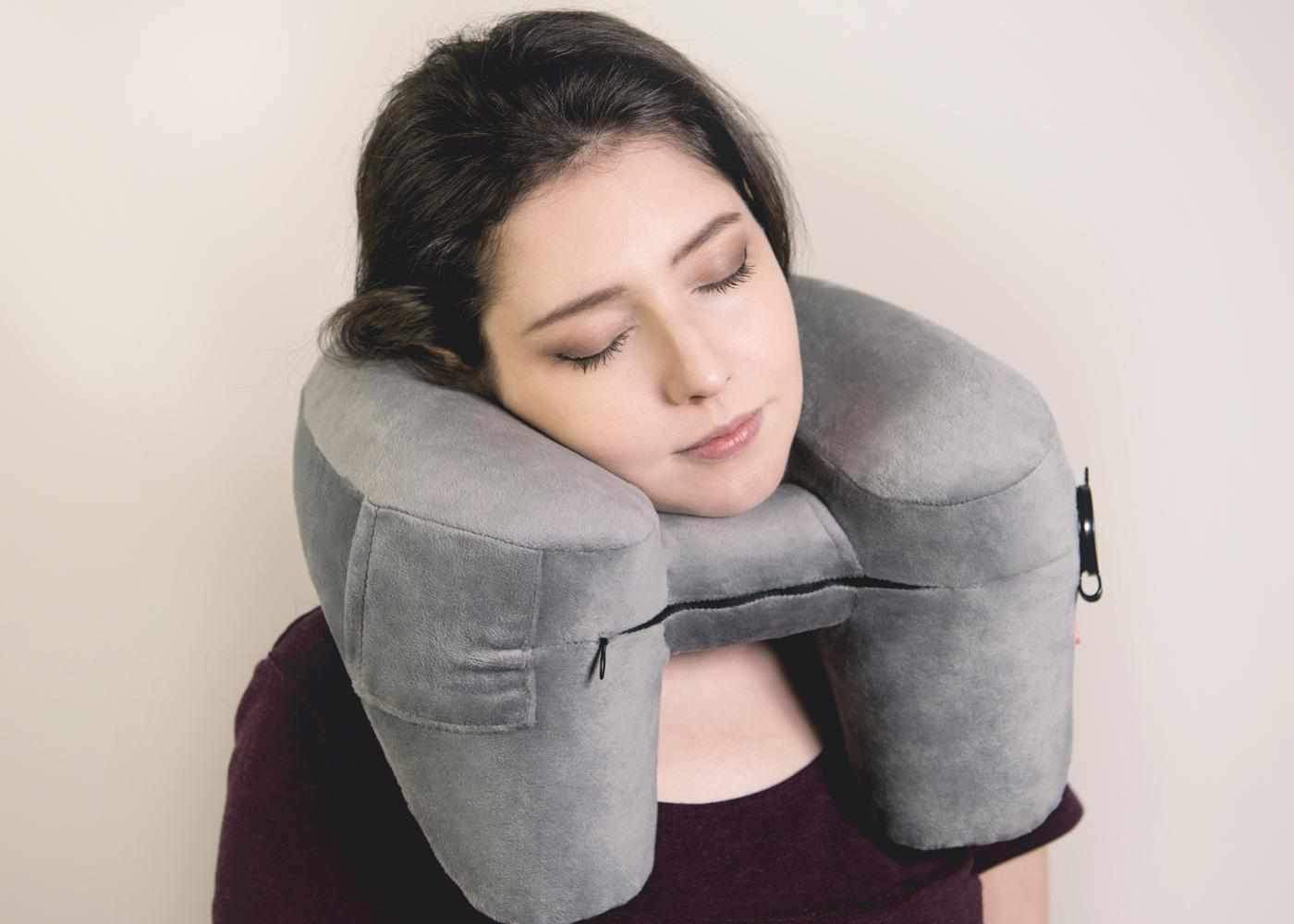 A woman resting comfortably with eyes closed, her head supported by a grey plush ergonomic Manta Travel Pillow.