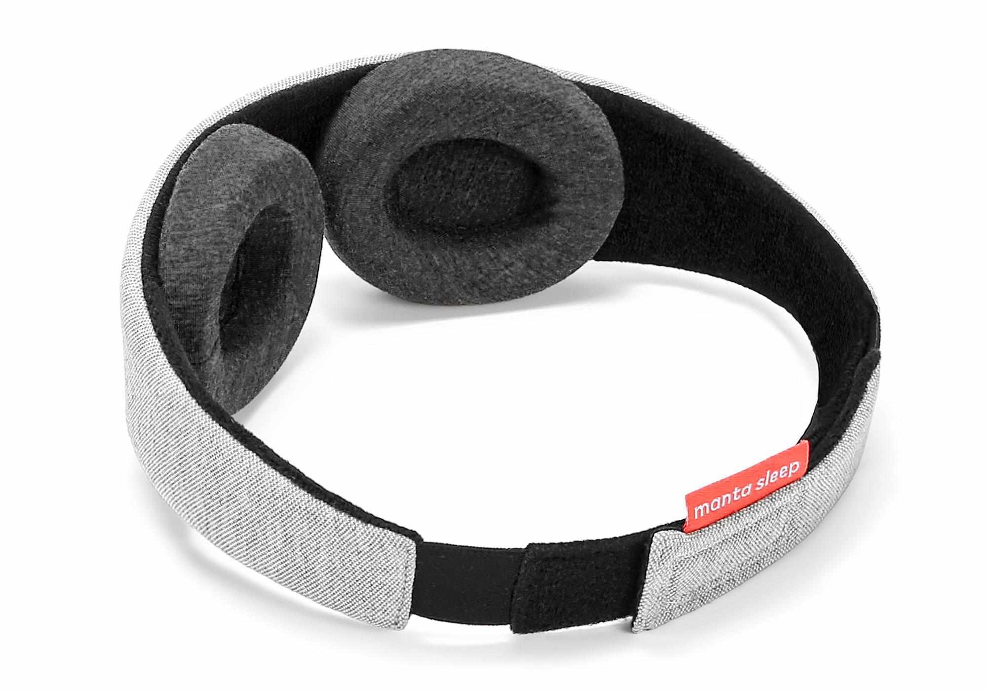 A three-quarter rear perspective of the Manta Sleep Mask isolated on a white background, showcasing the contoured shape of the dark gray plush eyecups on the black inner lining, the adjustable light gray heathered exterior strap, and the small coral-red brand label near the rear closure.