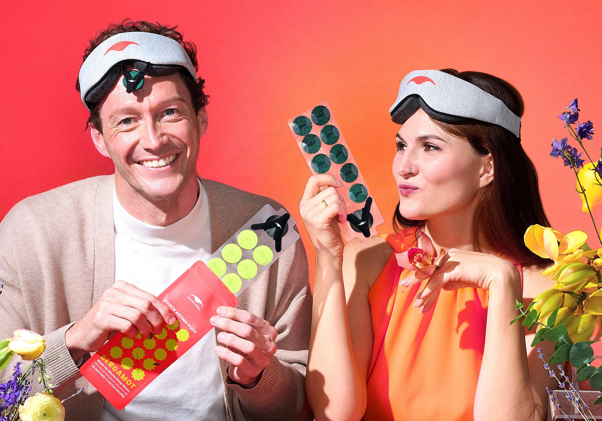Two people wearing sleep masks pull sheets of colorful aromatherapy patches out of their packets while posing playfully among vibrant flowers.