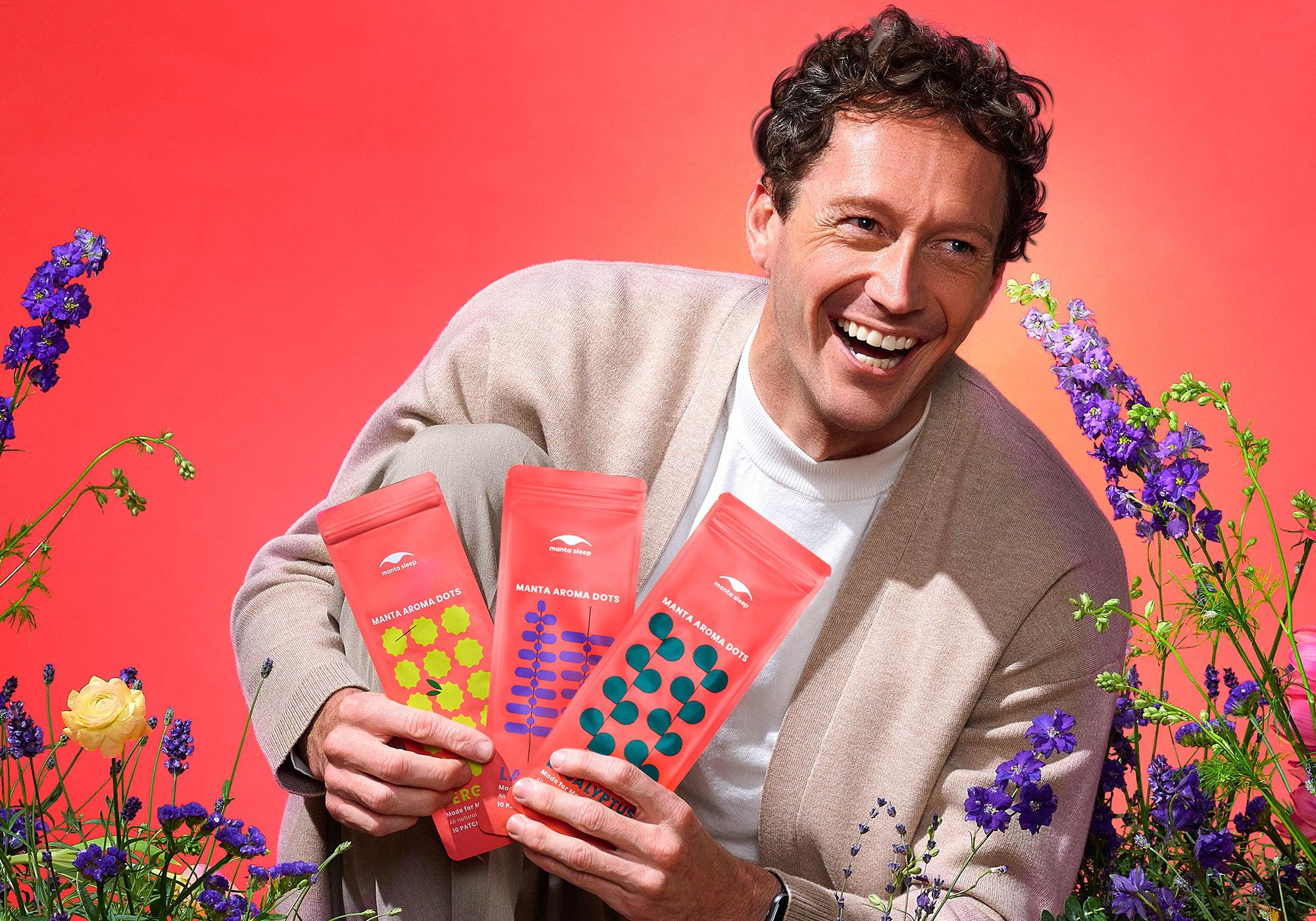 A smiling man holding three coral-red Manta Aroma Dots packets while sitting surrounded by colorful flowers, set against a bright coral background.