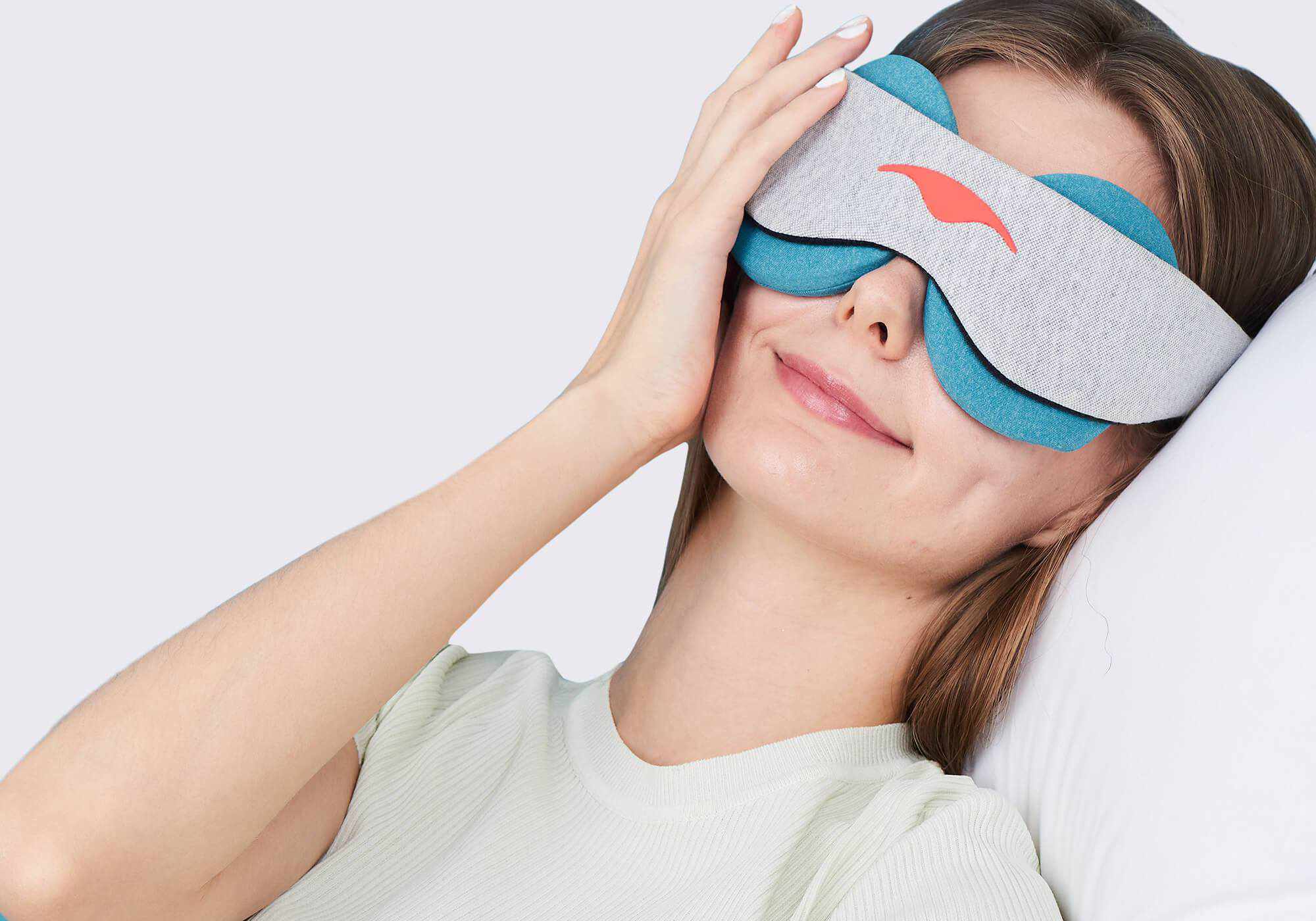 A woman resting her head on a white pillow while wearing a grey Manta COOL Sleep Mask fitted with blue-teal eye cups.