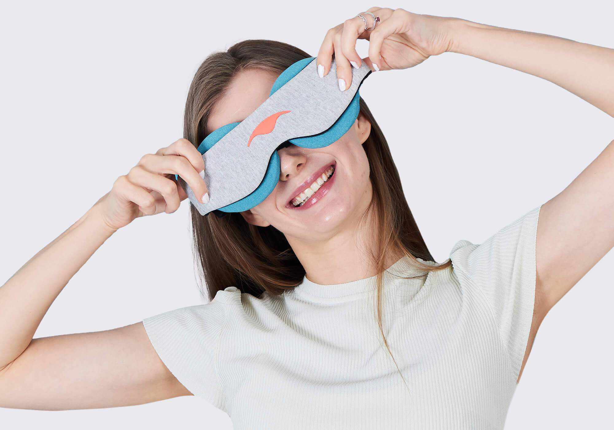 A smiling woman holding a grey Manta COOL Sleep Mask with a coral logo over her eyes, showing the blue-teal eye cups underneath, on a gray background.